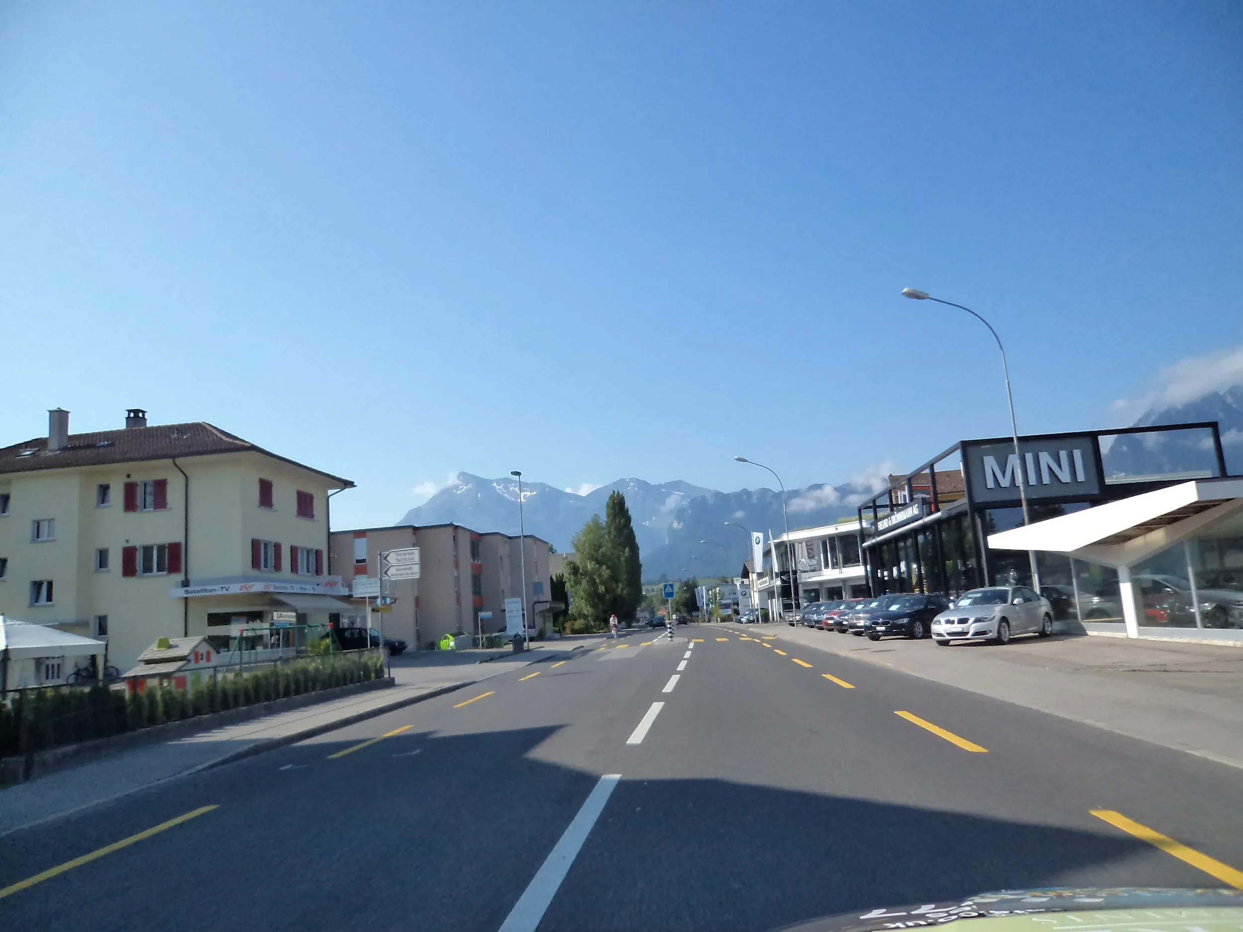 Street view with buildings, parked cars, and a mountainous backdrop under a clear blue sky.