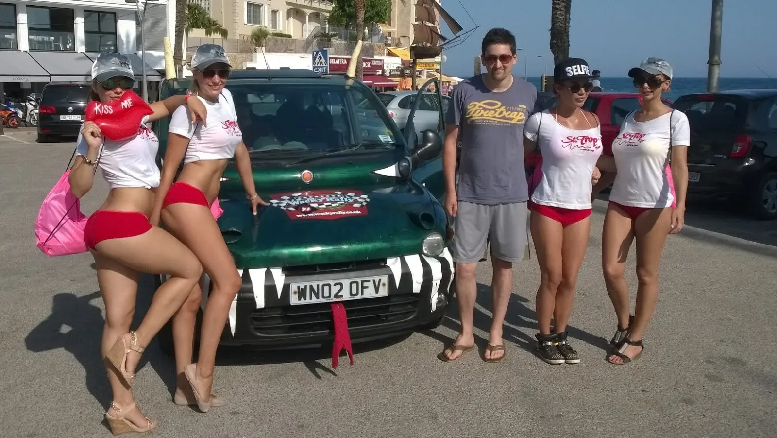 Group of five young adults posing in front of a small car on a sunny day at a beachside parking lot. Four women are dressed in white t-shirts and red shorts, with accessories like hats and sunglasses. One man wears a gray t-shirt and shorts. The back