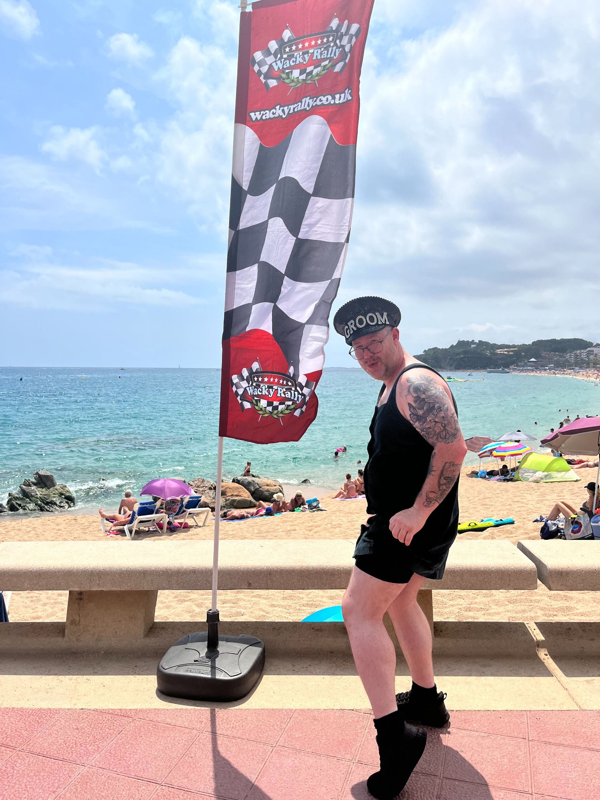 A man wearing a black tank top, shorts, and a hat labeled 'GROOM' posing on a seaside promenade. Behind him is a red and black checkered flag with the logo 'Wacky Rally' and a website link, near a beach with colorful umbrellas and people relaxing.