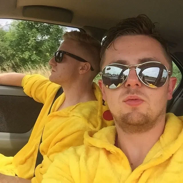 Two young men wearing yellow hoodies and sunglasses sitting in a car, with the one in front taking a selfie and the other looking out the window.