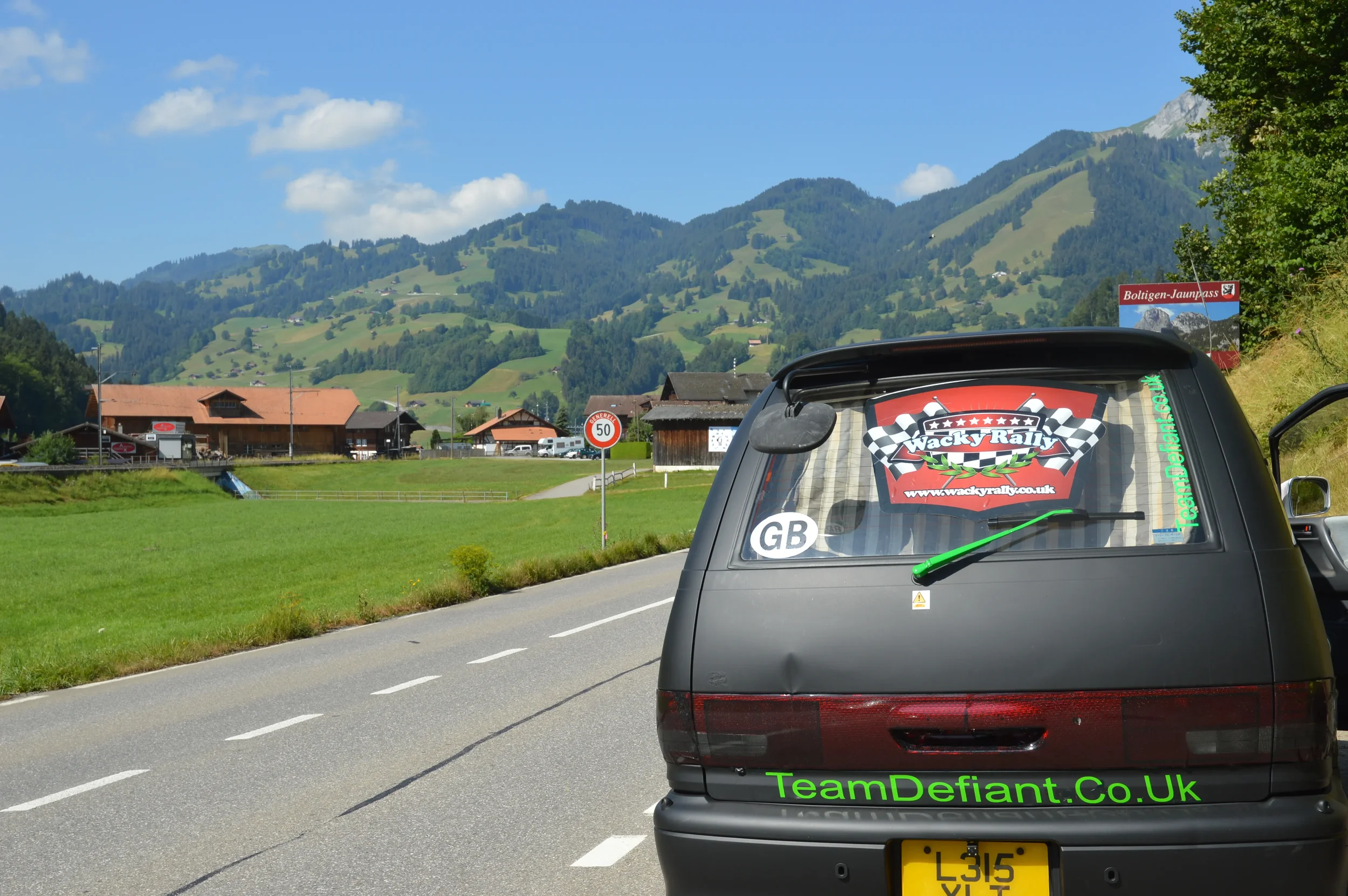 A black van parked on a rural roadside with stickers on the back, including one for 'Wacky Rally' and a green web address 'TeamDefiant.Co.Uk'. The background features rolling green hills, farm buildings, and a partly cloudy blue sky.