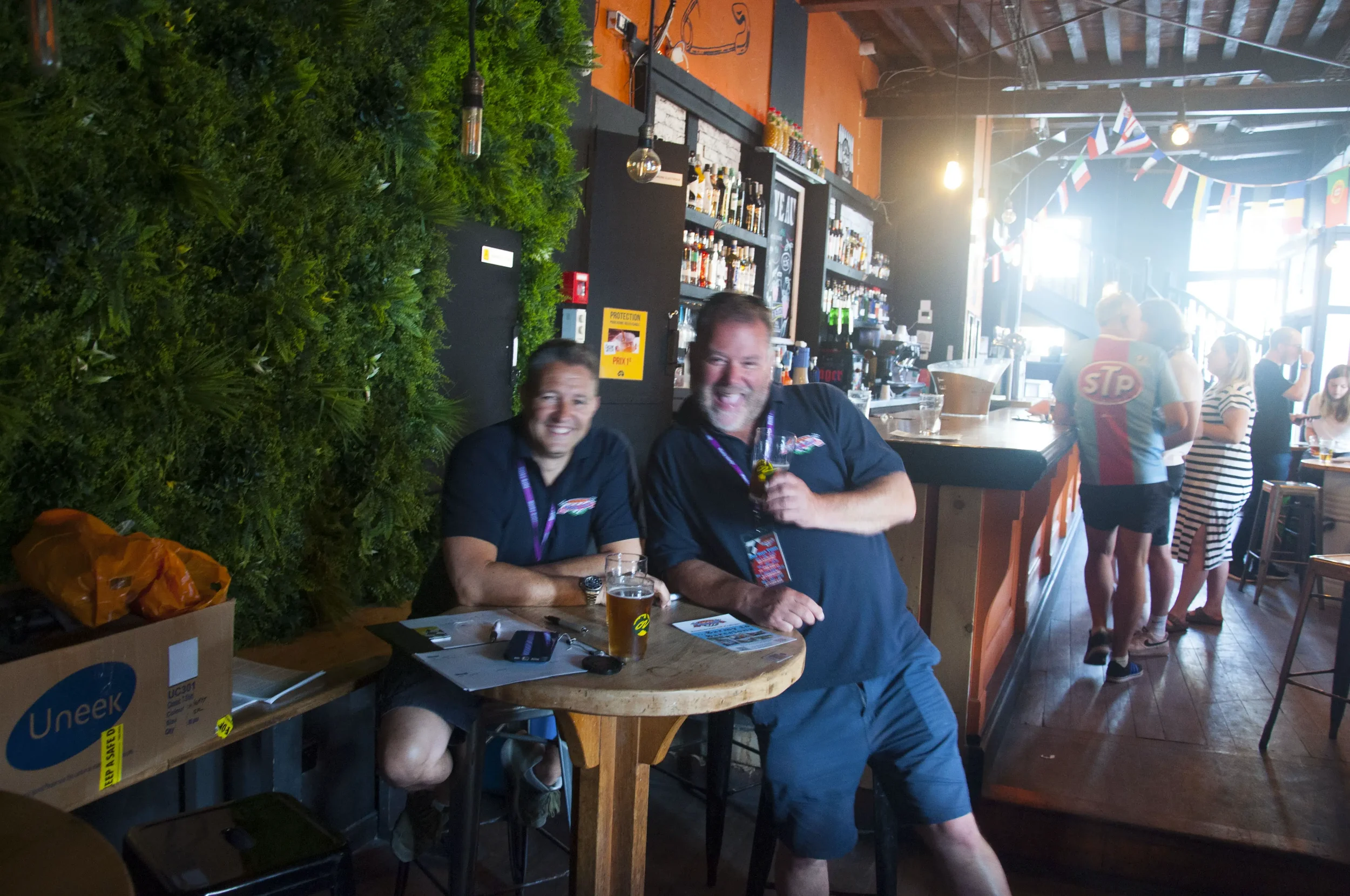 Two men sitting and standing at a small table inside a lively bar or restaurant, smiling and holding drinks, with a bar counter and several people in the background, and a lush green wall on the left side.