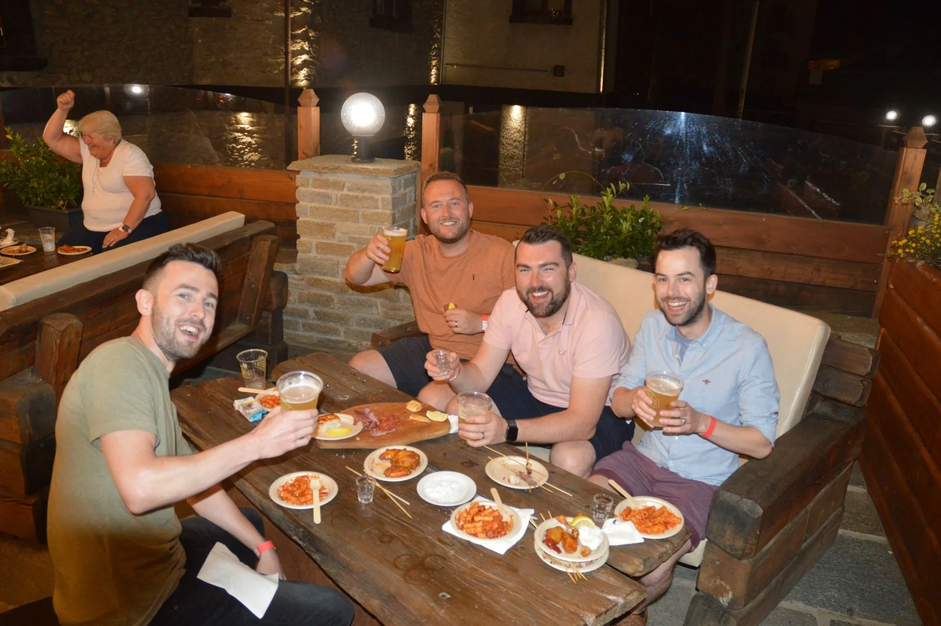 Four men sitting around a wooden table outdoors at night, enjoying drinks and appetizers, with additional people and a woman in the background. They are smiling and celebrating.