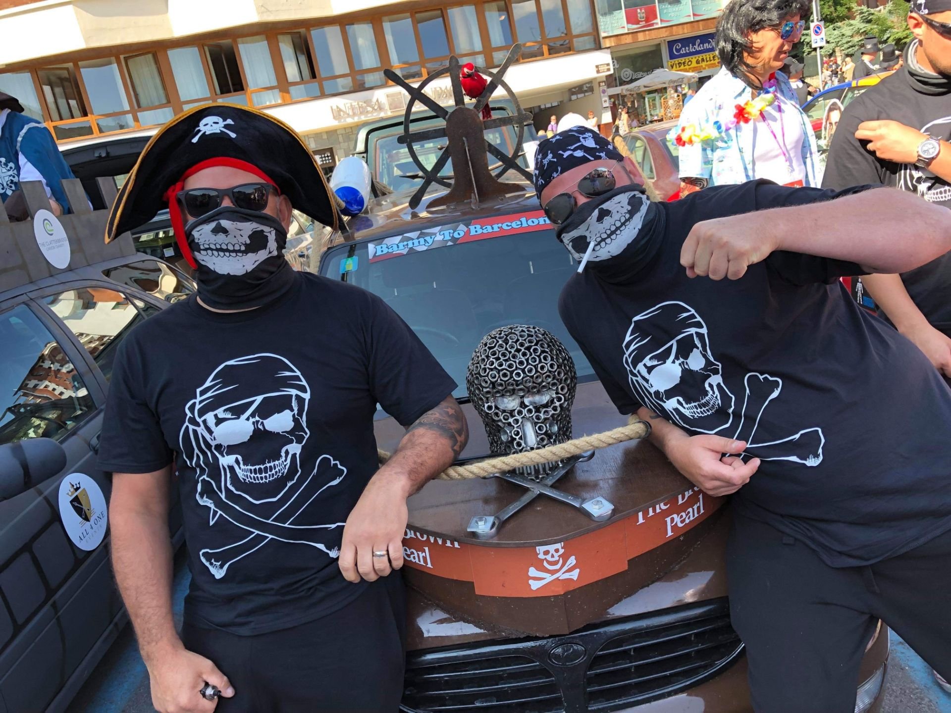 Two men dressed in pirate costumes with skull masks and pirate hats, standing next to a decorated car with a skull on the front and a sign that says "Barrys to Barcelona" in a busy outdoor setting.
