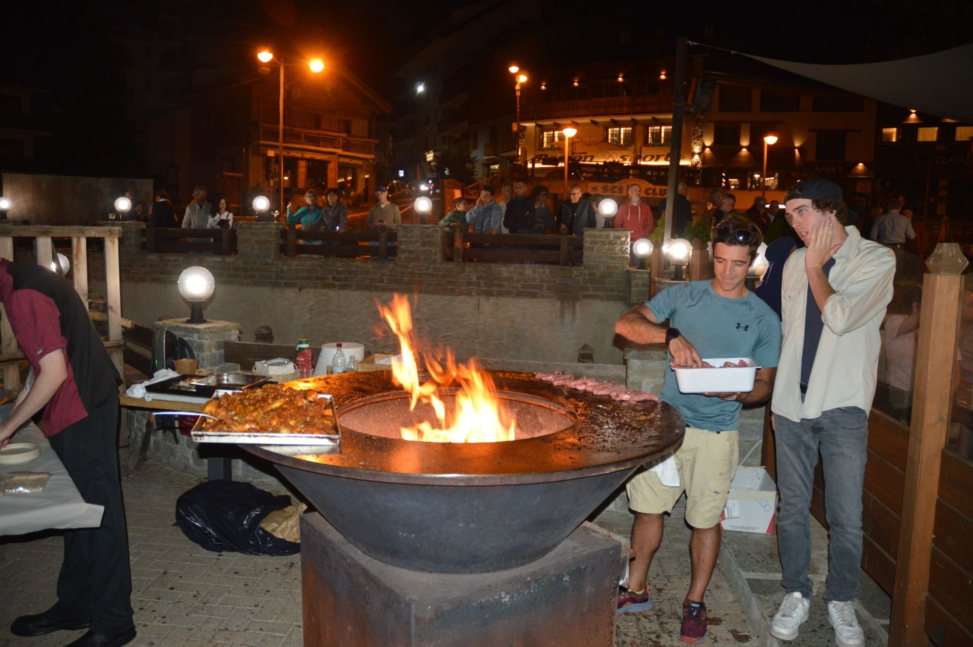 Two young men preparing meat at a night outdoor barbecue with a large fire pit, with a crowd of people on a bridge in the background.