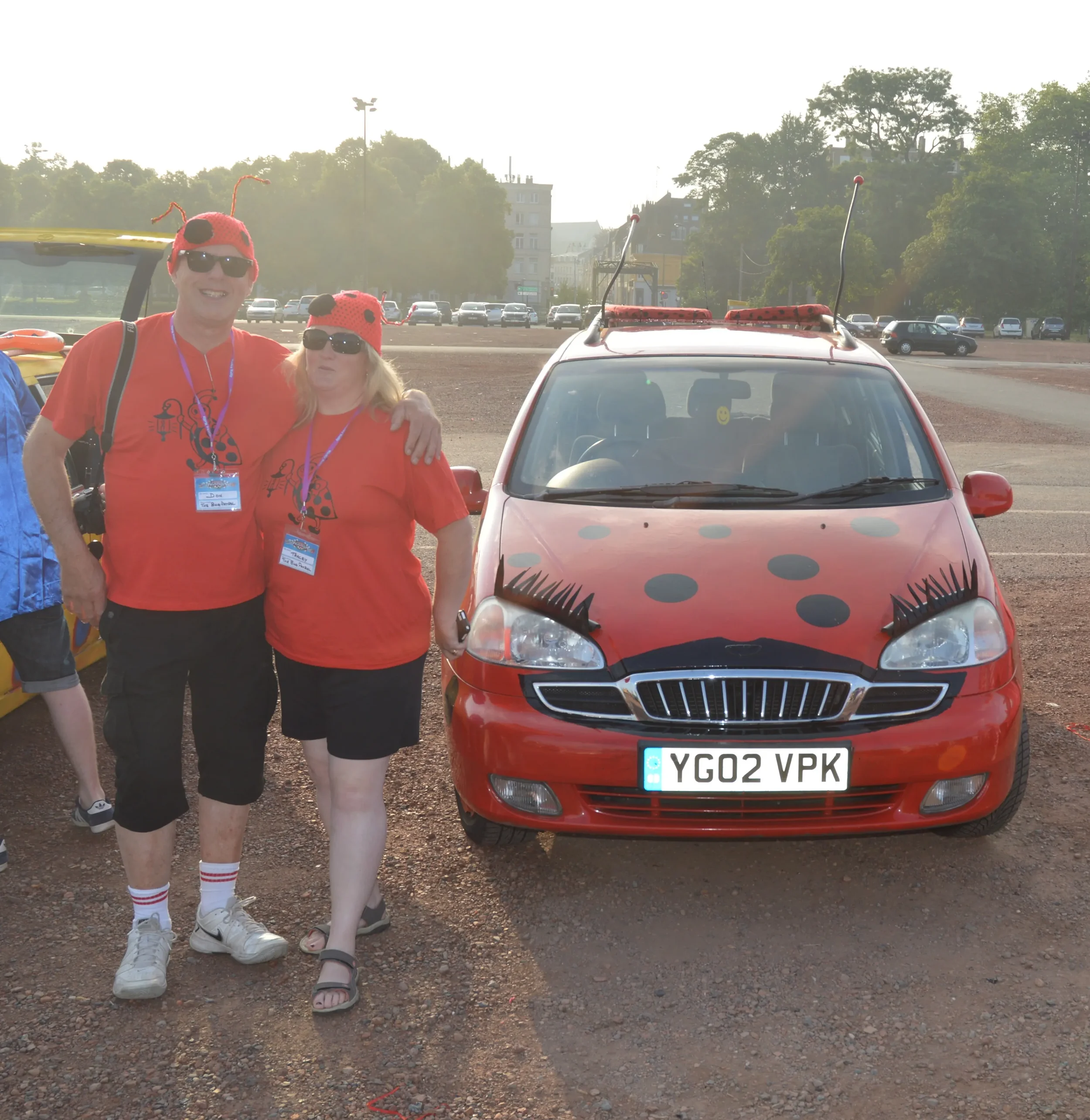 Two people wearing ladybug costumes with red shirts, black pants, and ladybug hats, standing beside a decorated red car with ladybug and eyelash designs, in a parking lot on a sunny day.