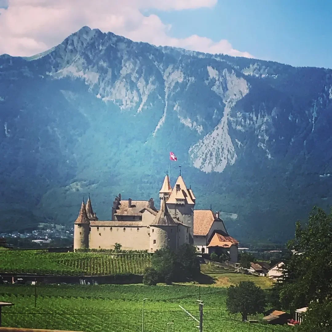 A castle with a Swiss flag, set against a backdrop of mountains and vineyards, under a partly cloudy sky.