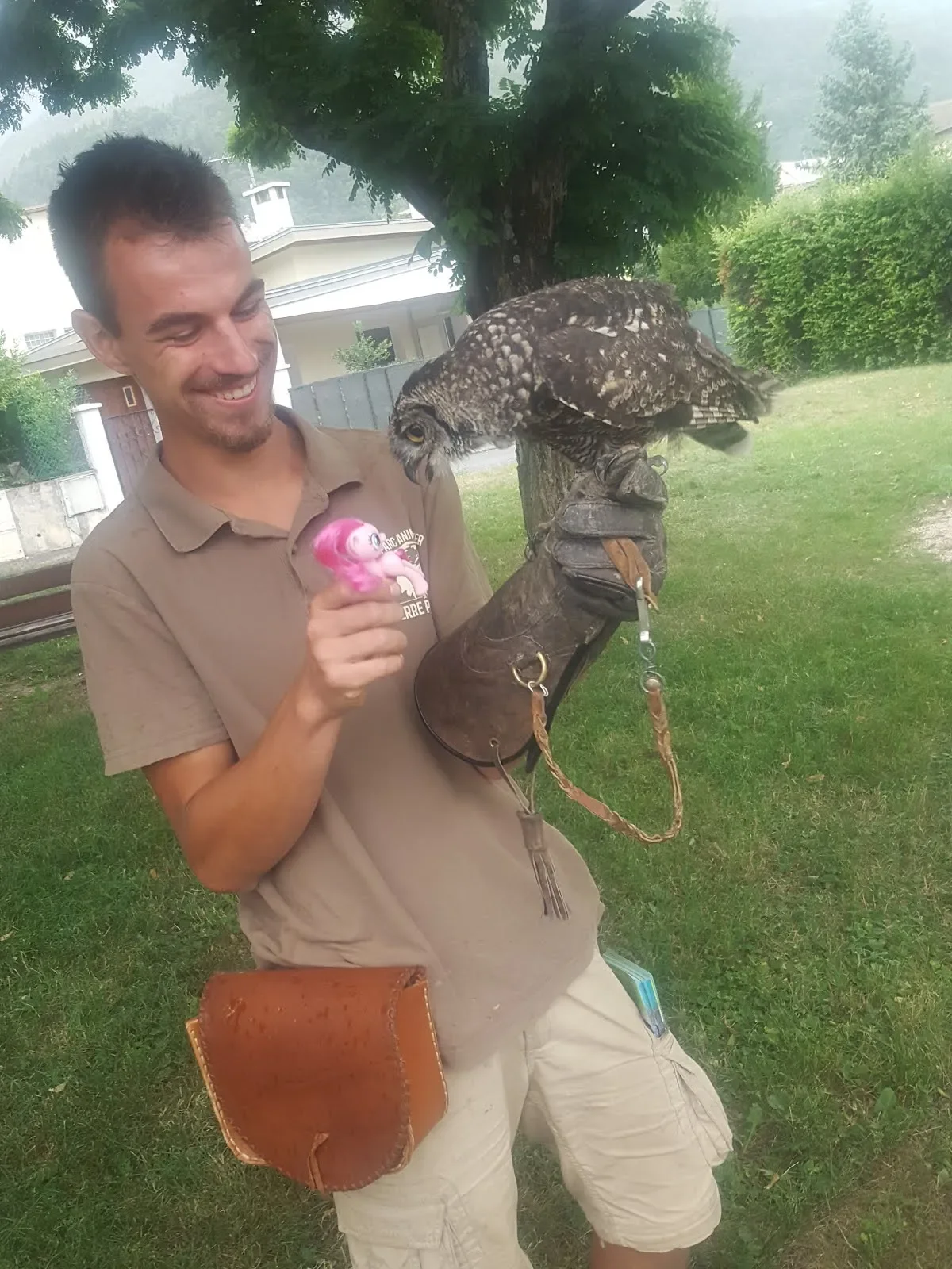 A young man smiling as he holds onto a large owl on his gloved arm, in an outdoor park setting with trees and grass in the background.