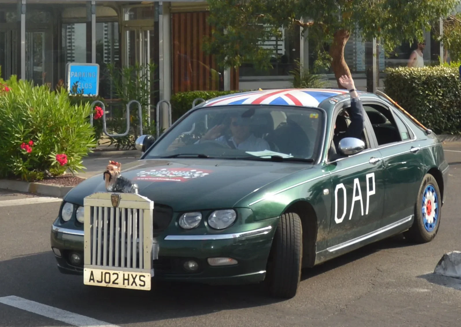 A green car with the word 'DAP' on the side, decorated with Union Jack patterns on the roof. There is a small animal figure on the front grille of the car and a person inside waving out the window. The car is parked near a building with bushes and a 