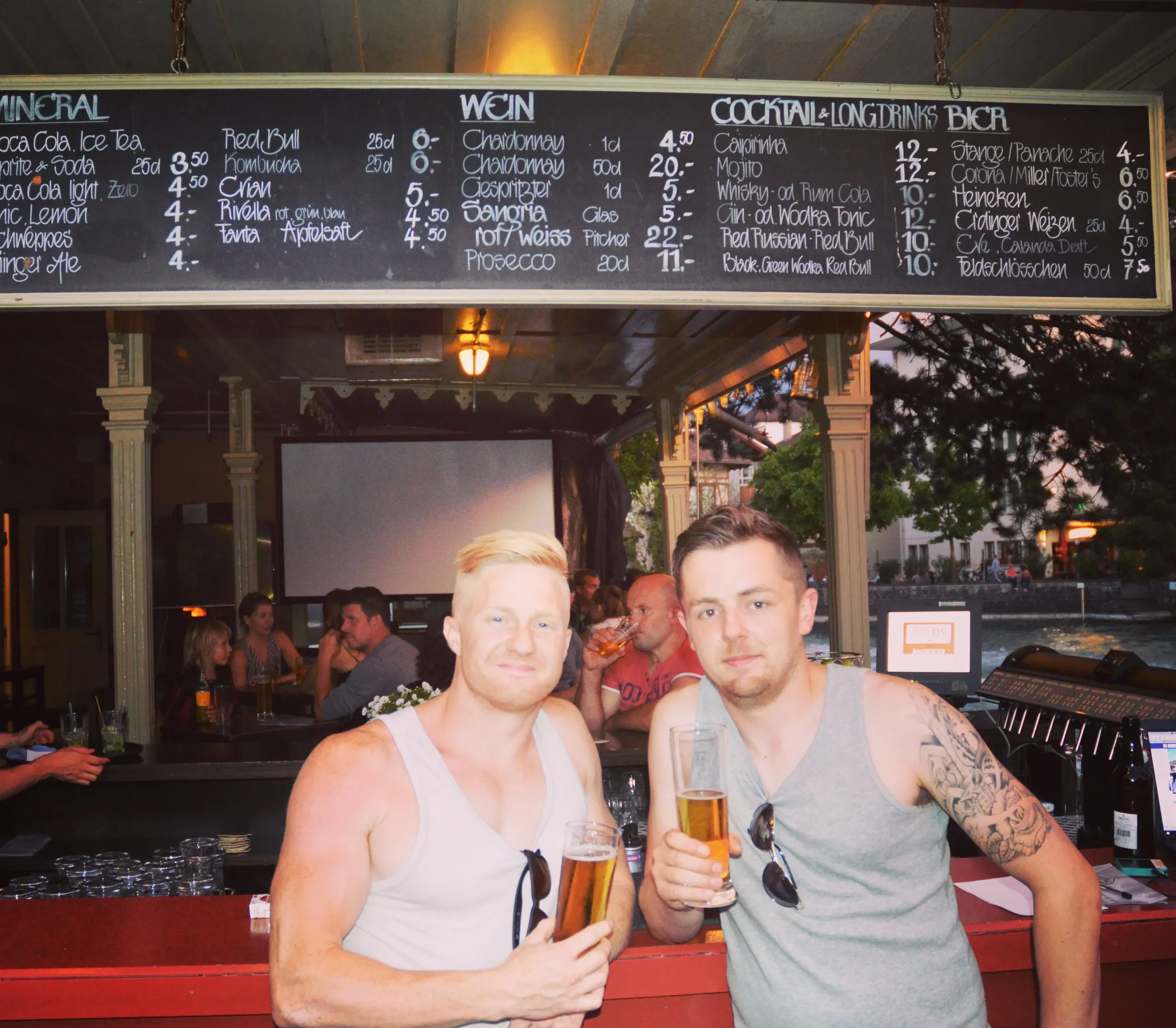 Two young men in sleeveless shirts holding glasses of beer at a bar with a chalkboard menu above them; other patrons seated in the background, with outdoor trees and a waterway visible outside.