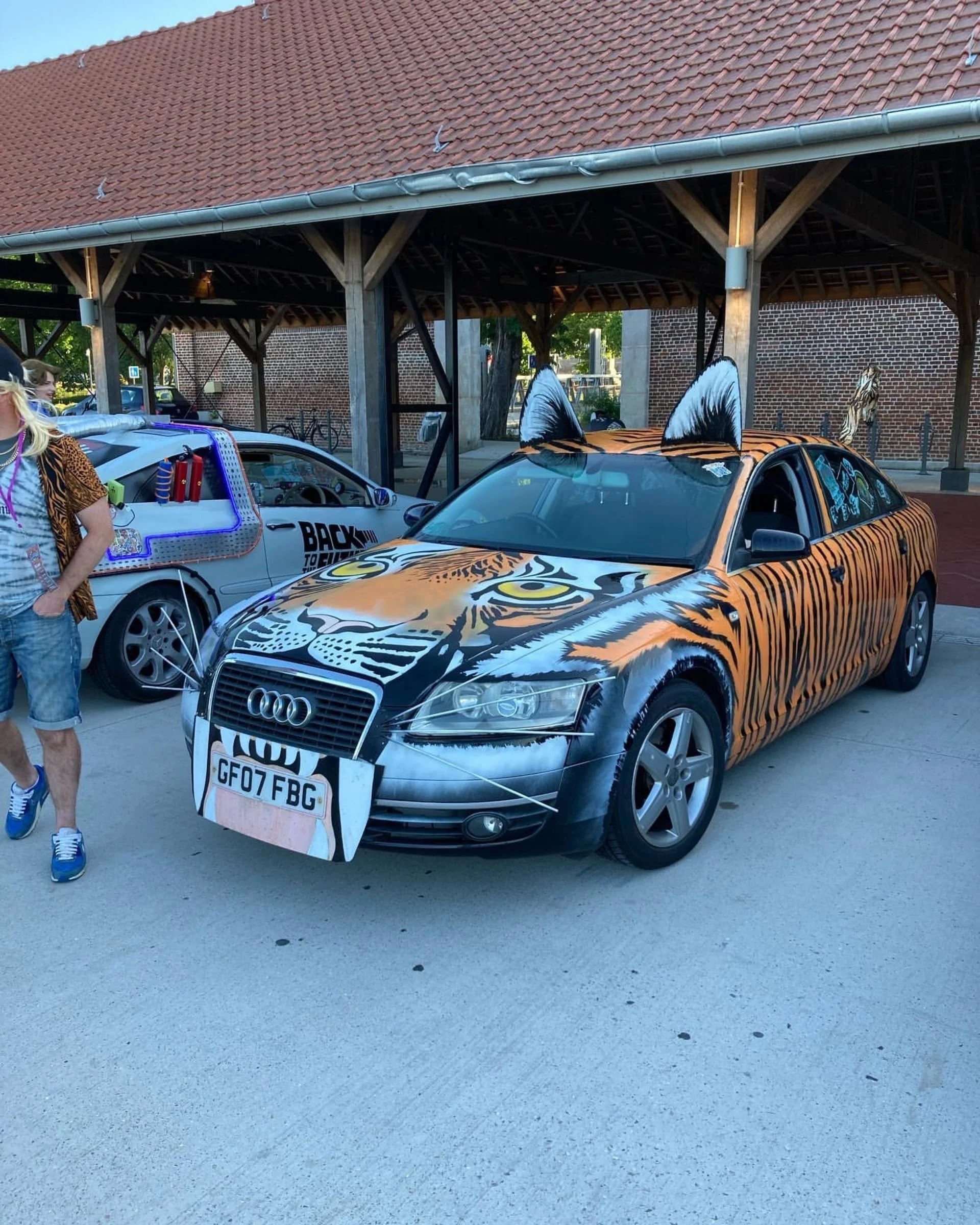 A black Audi car decorated with a tiger face and stripes in orange, black, and white, with tiger ears on the roof and tiger face painted on the front, parked under a wooden shelter next to another decorated vehicle.