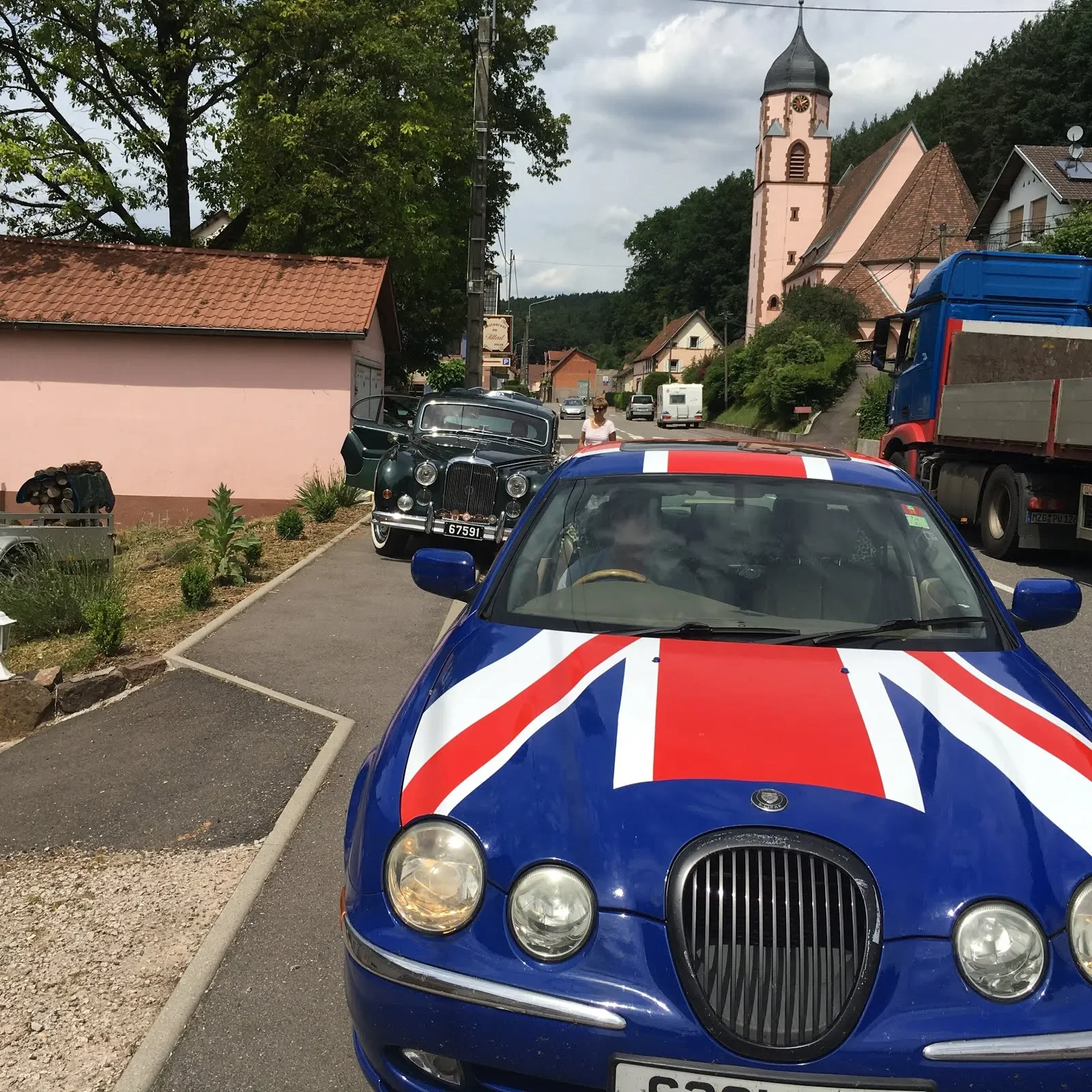 A street scene with a blue car featuring a Union Jack design parked in the foreground, an older black car in the background, and a church with a tall steeple on a hilly street in a small town surrounded by trees.