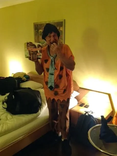 Person dressed as Fred Flintstone eating snacks in a hotel room.