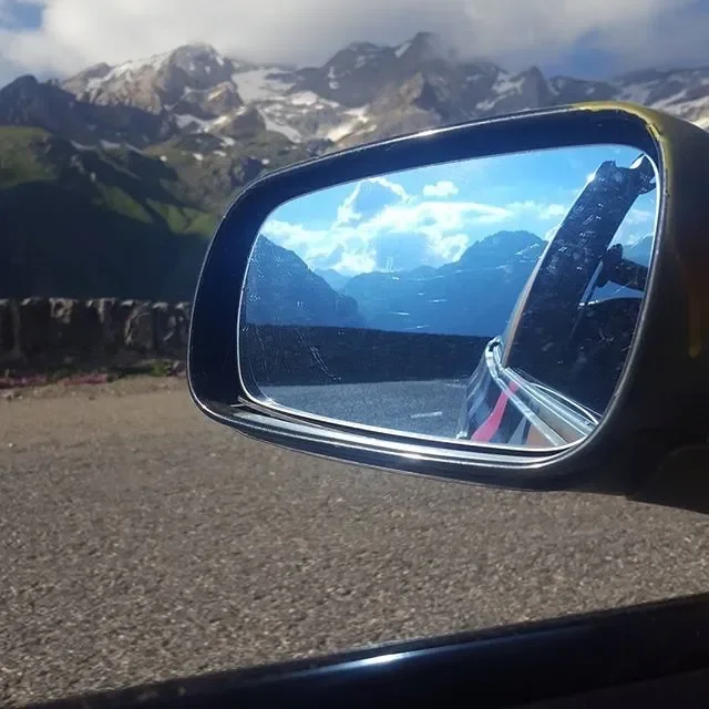 View of snow-capped mountains and blue sky reflected in a car's side mirror, with a mountain road in the foreground.