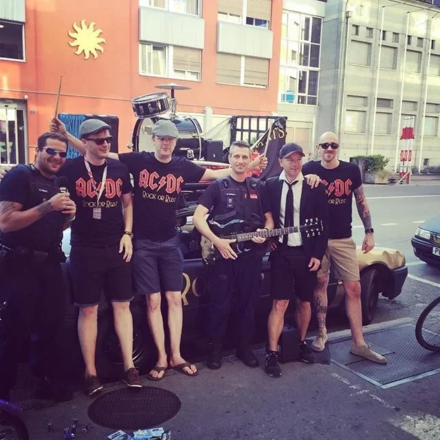 Six men standing on the street in front of a vehicle with a drum set on it, some wearing AC/DC band shirts, holding drinks and musical instruments, with buildings in the background.
