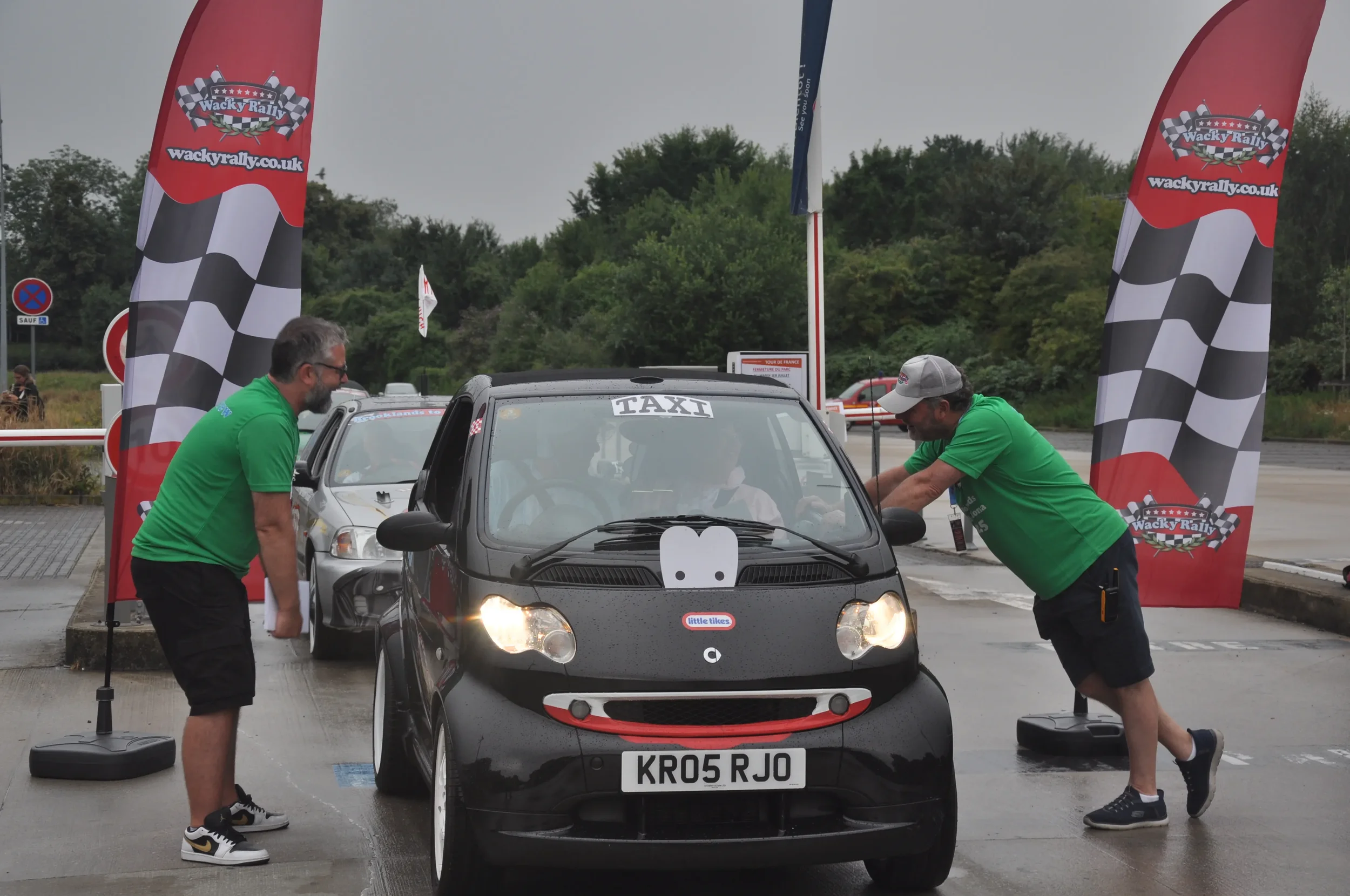 Two men in green shirts interacting with a small black car at a rally event, with checkered flags and banners in the background.