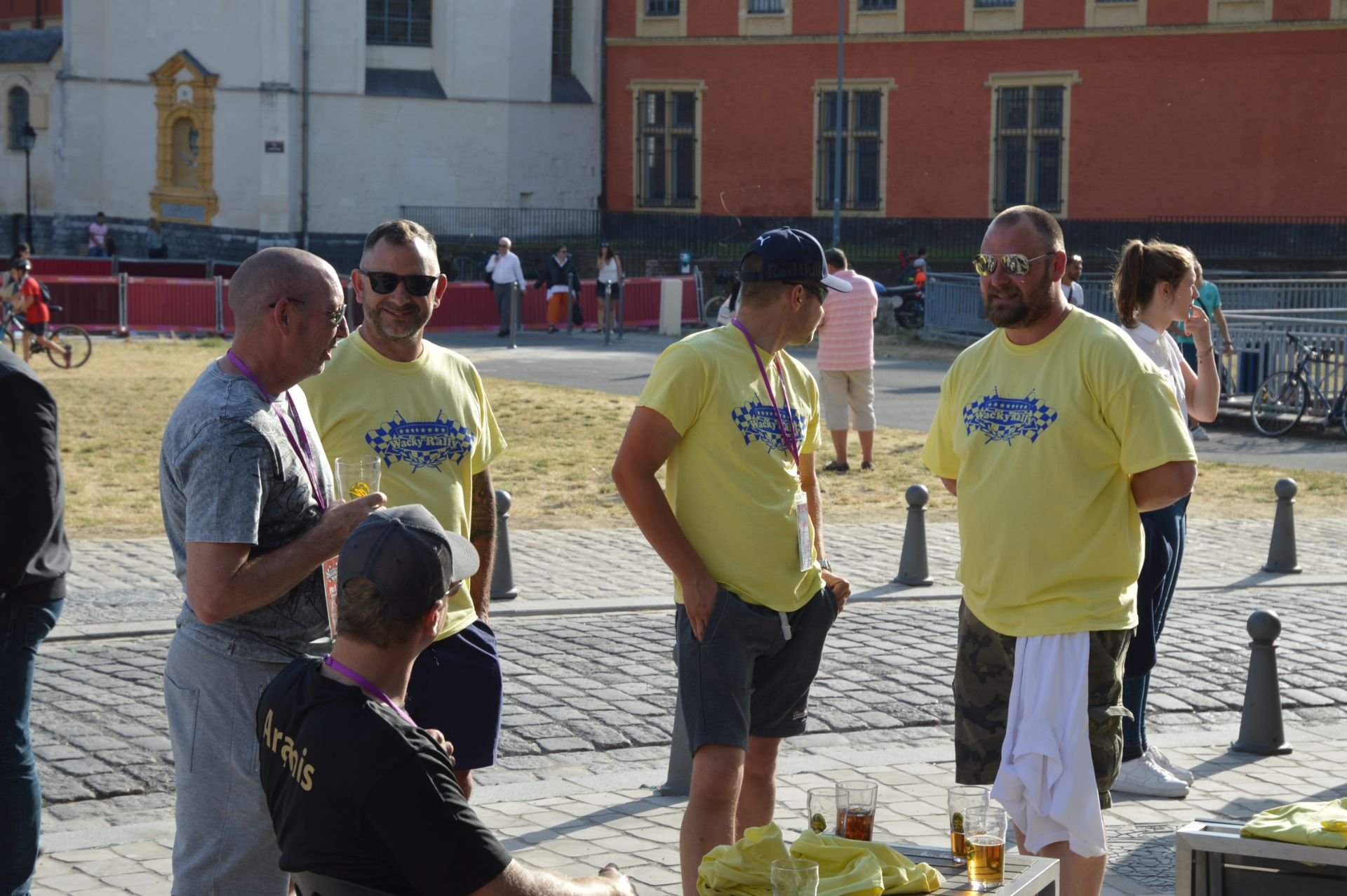 Group of men and women standing and sitting outdoors near a table with drinks, some wearing yellow T-shirts with a checkered logo, in a city setting.