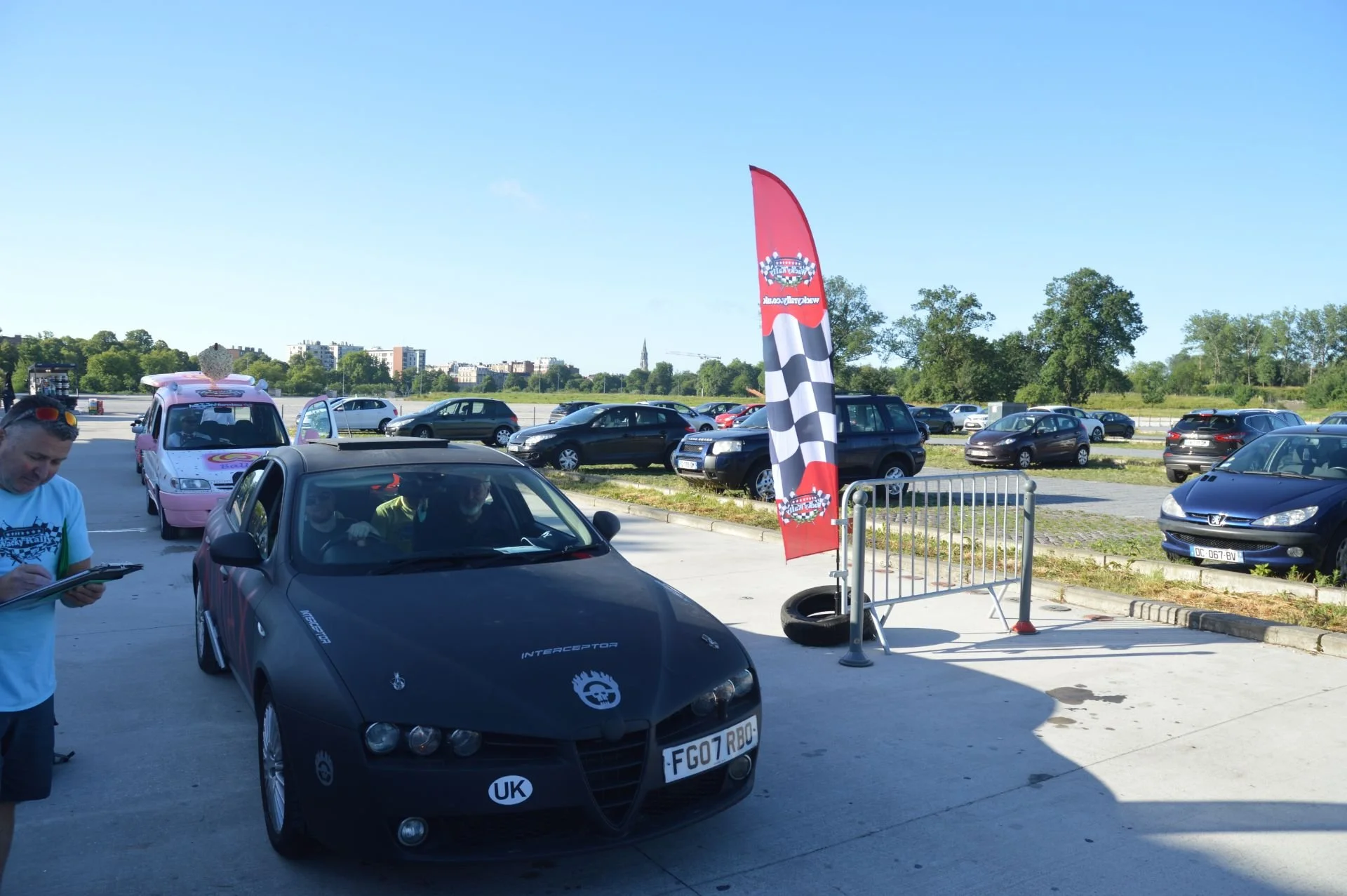 A black sports car with decals, parked under a racing-themed flag, with a person standing nearby writing on a clipboard and other cars in the background.