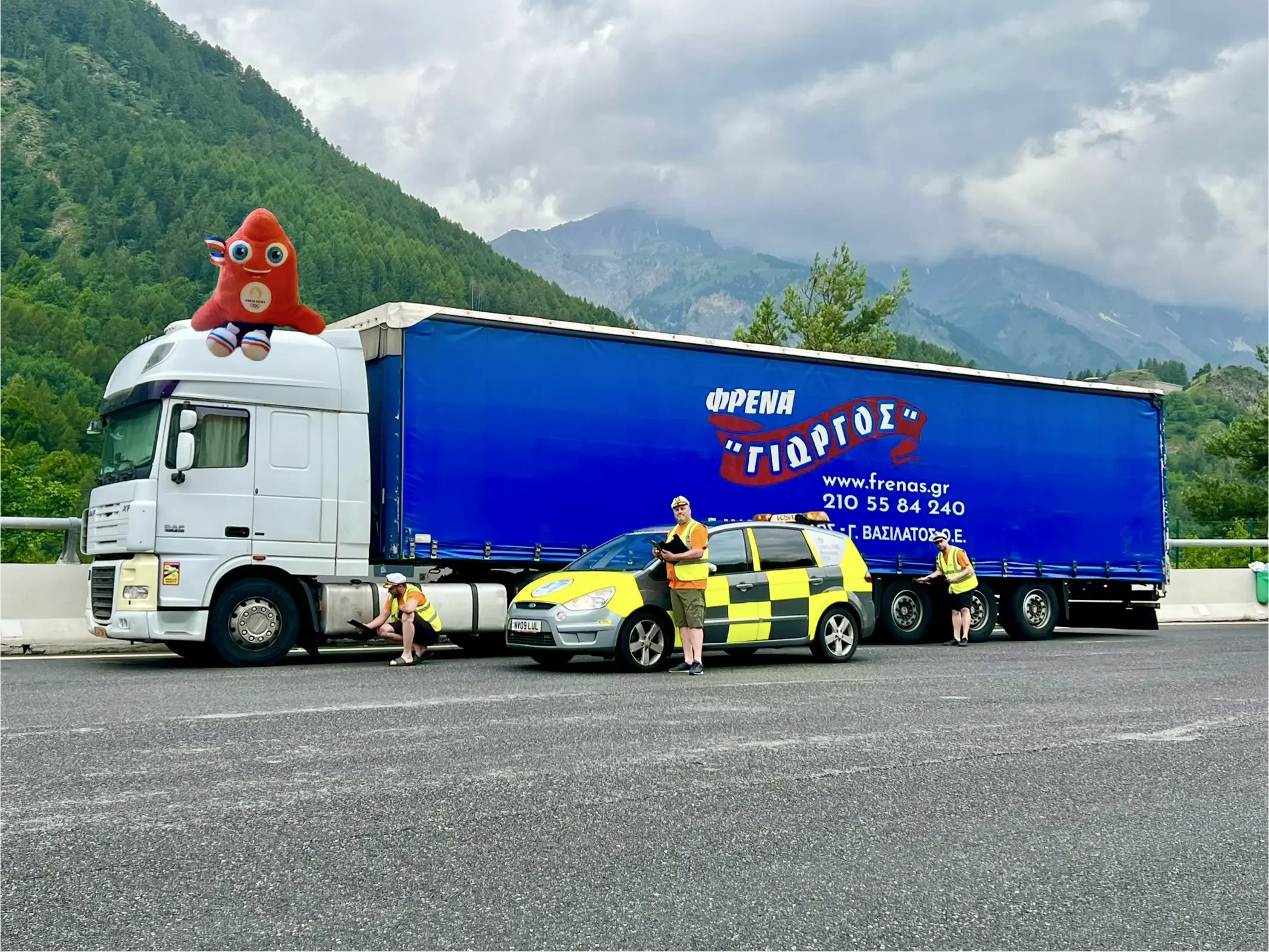A large white semi-truck with a blue trailer, featuring Greek writing and contact information, parked on a highway with mountains and cloudy sky in the background. Three workers in yellow safety vests are attending to the truck, and a police car is p