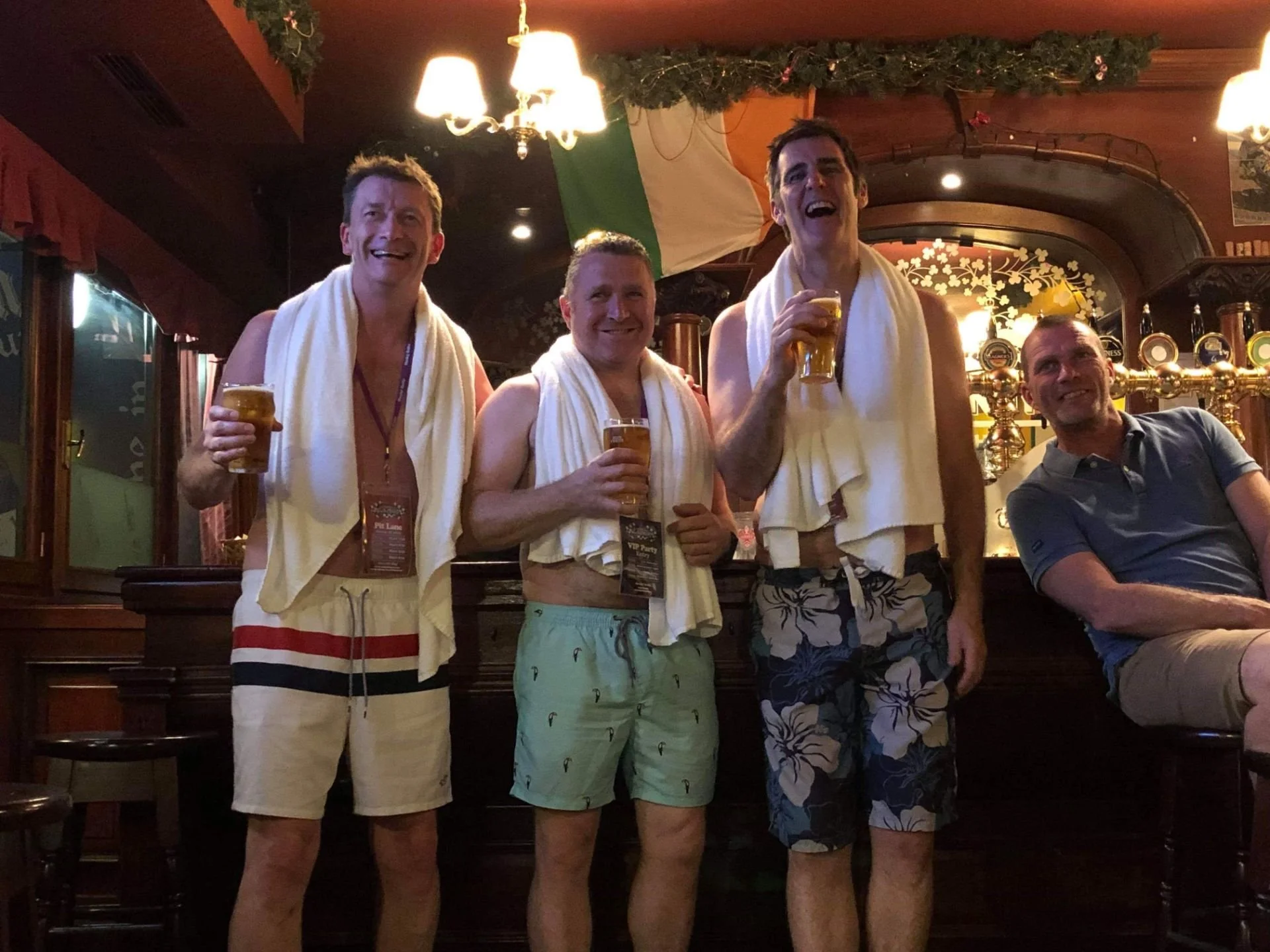 Four men in a pub celebrating, holding beers, with bathing towels draped around their necks, some wearing swim shorts, and smiling.