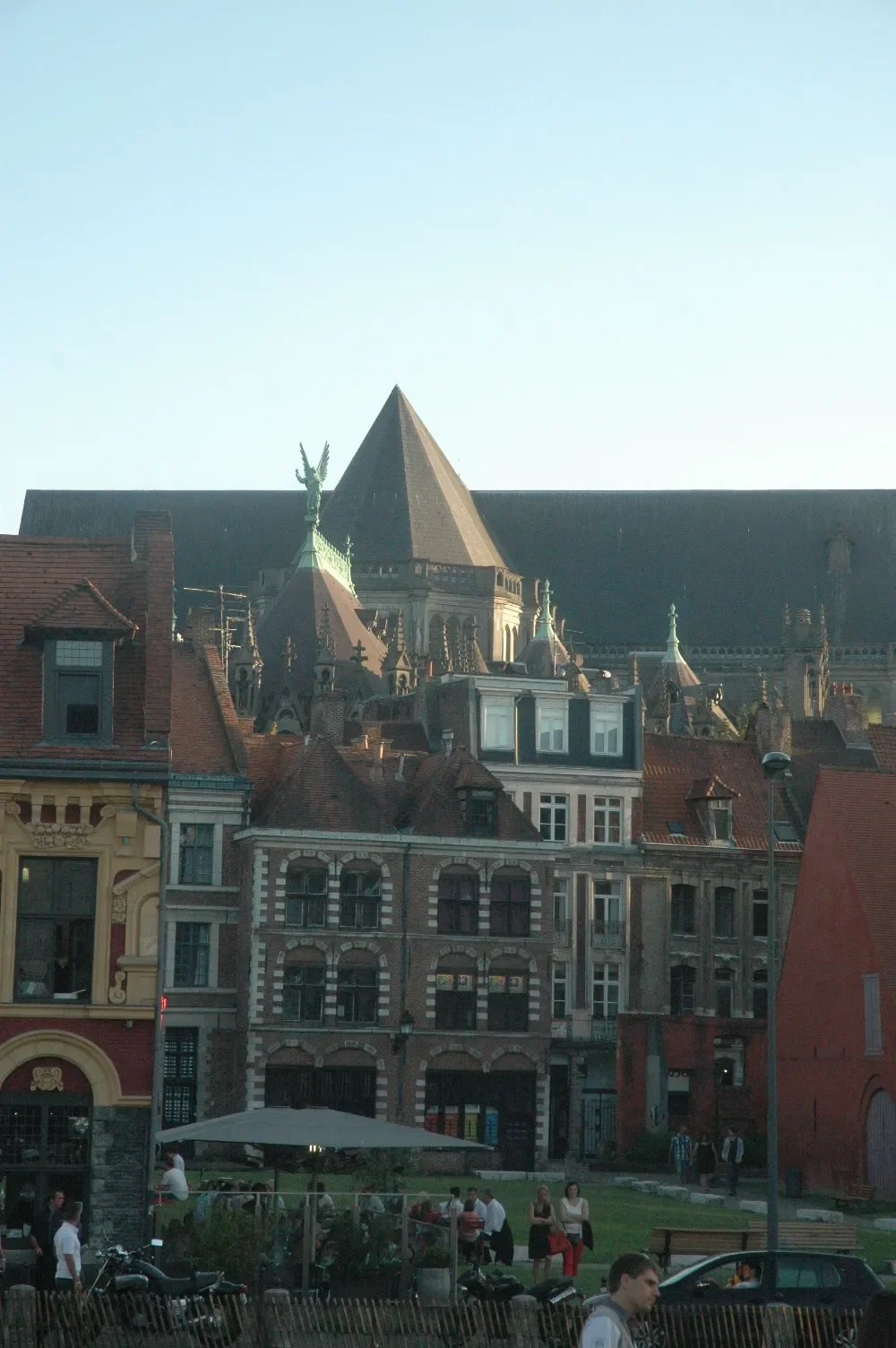 European city street with historic buildings, people, and parked motorcycles in the foreground, and a church with a pointed roof and decorative statues in the background.