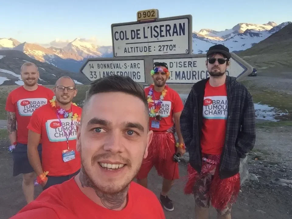 Group of five men dressed in red t-shirts and Hawaiian leis posing for a selfie at Col de L'Iseran mountain pass with snow-covered mountains in the background.