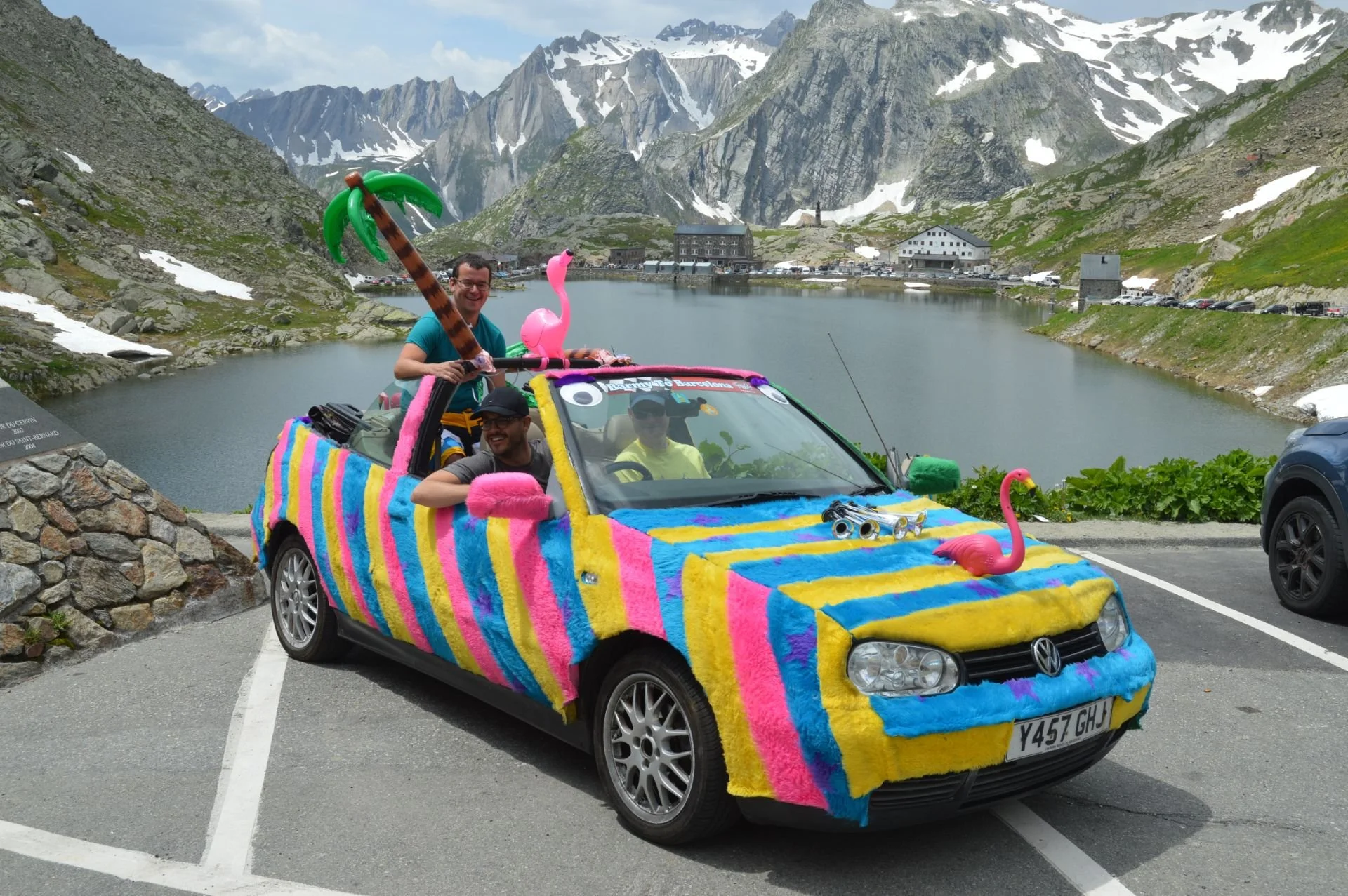 Colorfully decorated Volkswagen car with pink flamingo and inflatable palm tree accessories parked near a mountain lake, with three men inside smiling, overlooking snowy mountain peaks.