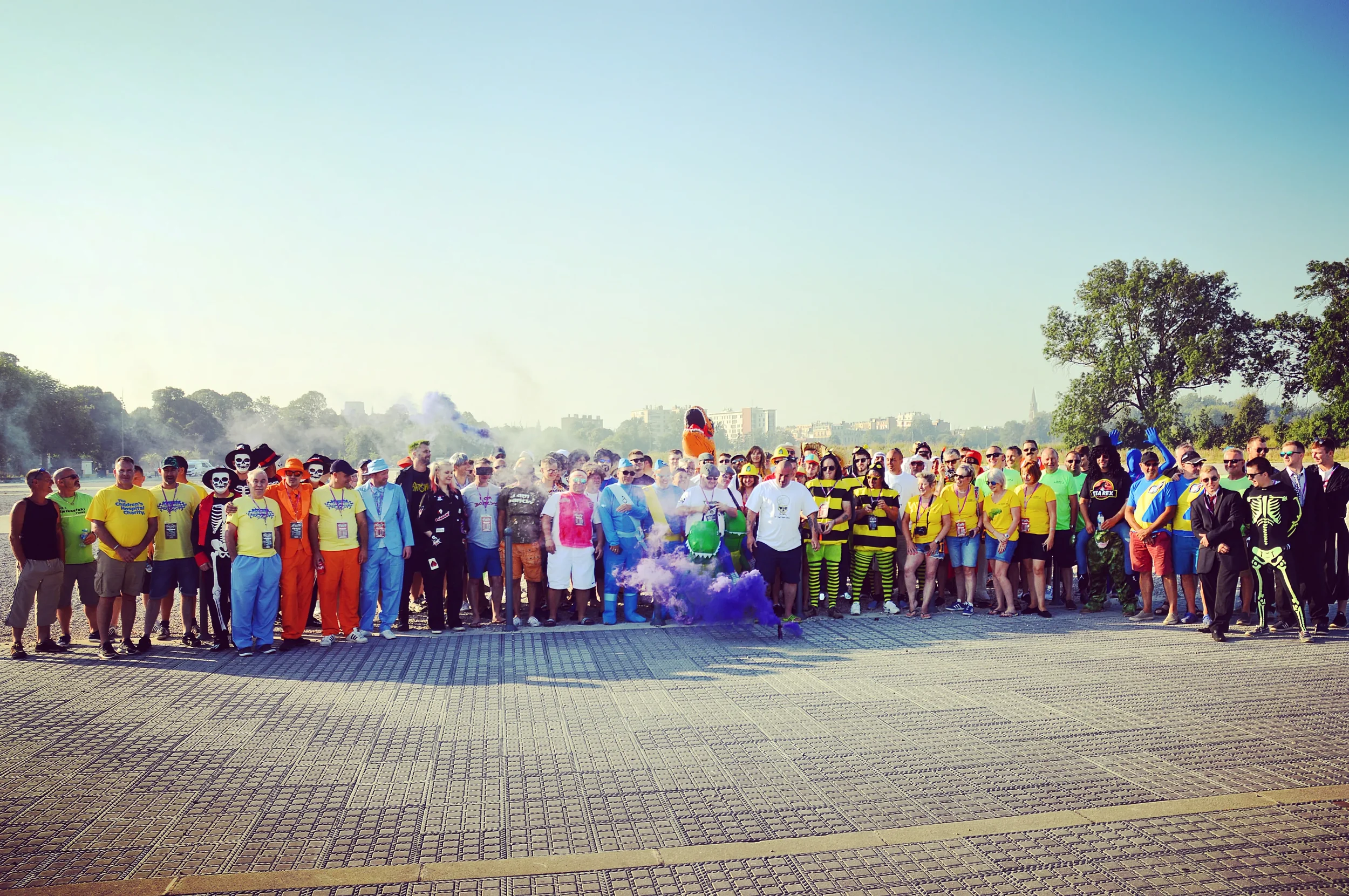 Large group of people dressed in colorful costumes, some in skeleton, bee, and other themed outfits, gathered outdoors on a sunny day for a charity walk or event. Many are wearing badges or medals, standing on a paved area with trees and cityscape in