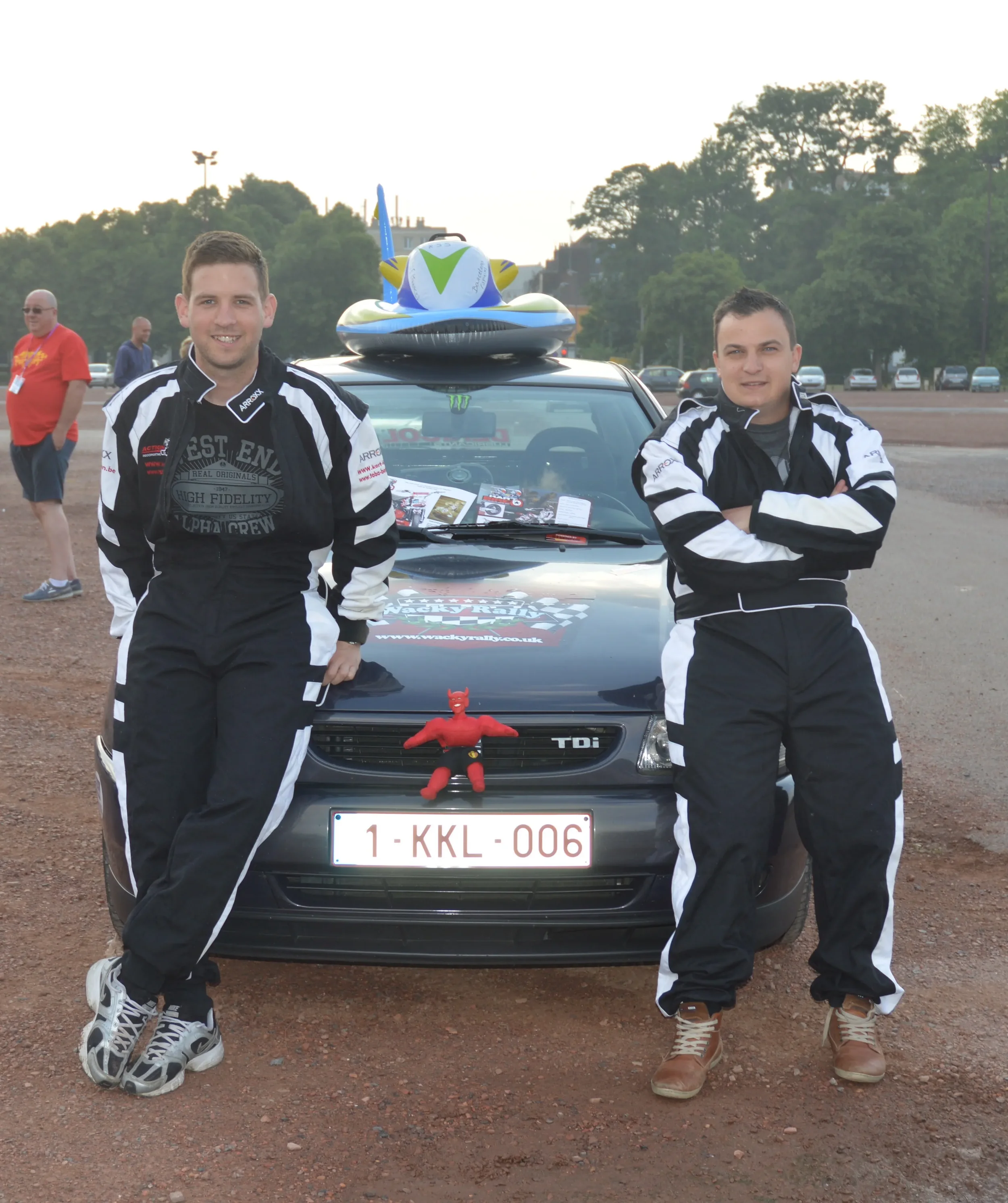 Two men in racing suits standing next to a black racing car with Belgian license plate, with a racing toy devil and a bobblehead on the front grille, and a dog-shaped float on the roof. They are at an outdoor event with people and trees in the backgr