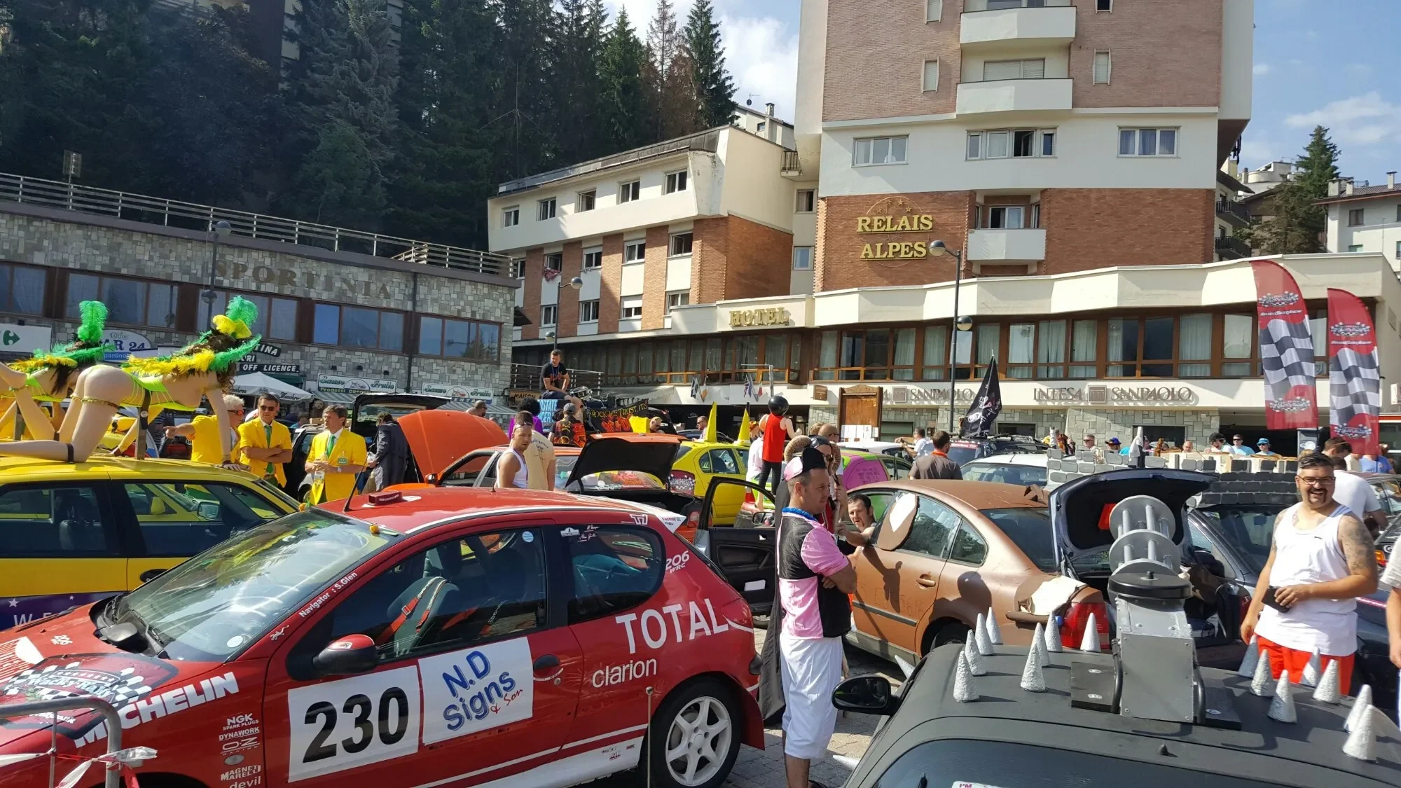 The image shows a lively outdoor event with numerous custom cars, people walking around, some dressed in costumes, and flags. In the background, there is a building labeled 'Relais Alpes' and a hotel. The scene appears to be a car show or rally in a 