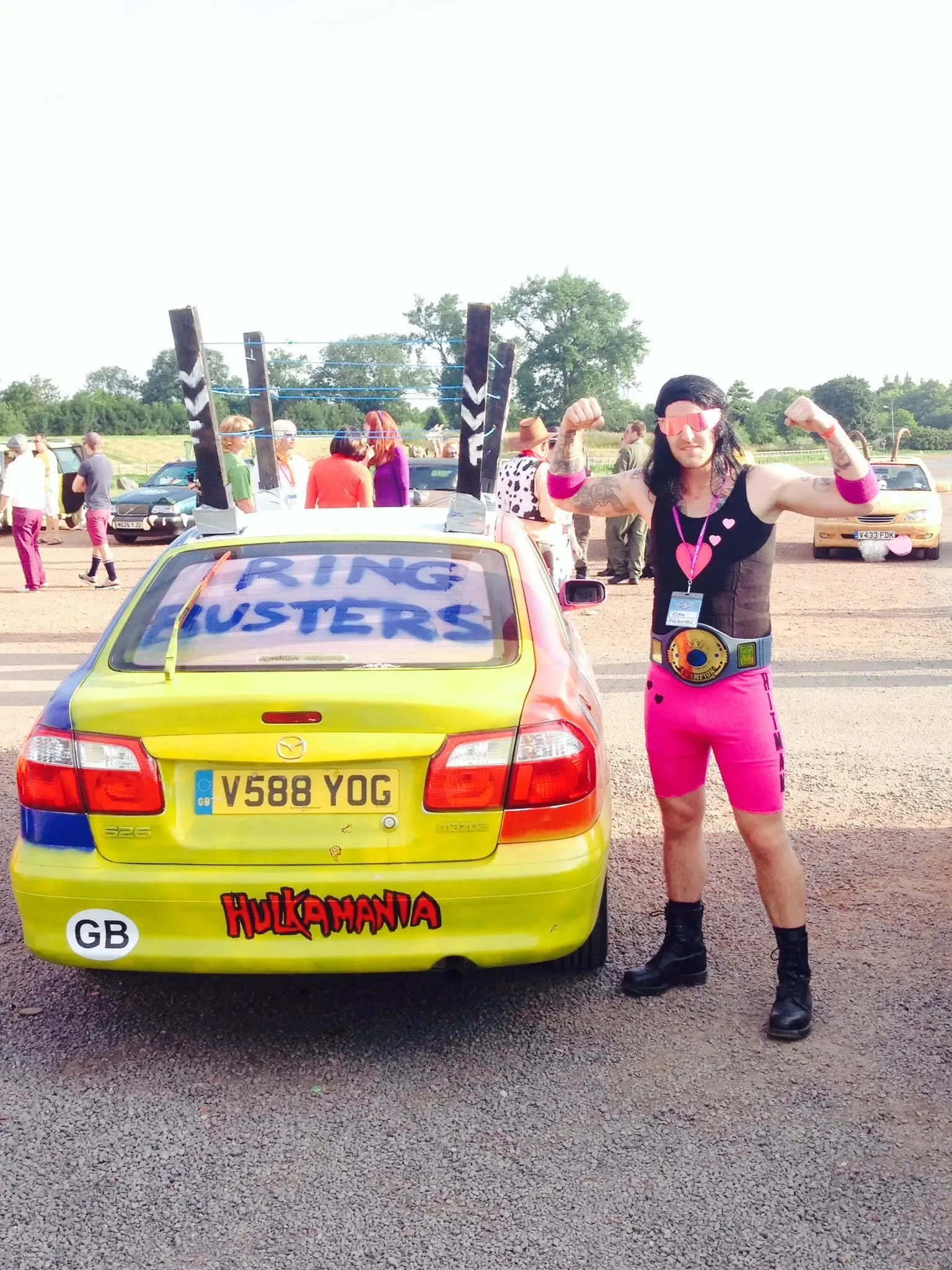 Person dressed as a wrestler posing with a car at an outdoor event, with people in the background and a sign on the car that says "Ring Busters".