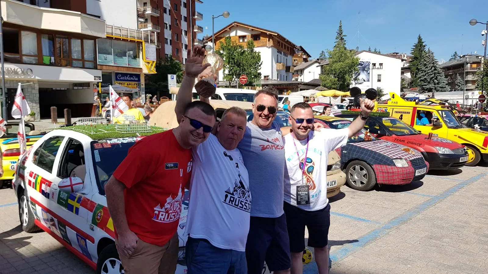 Four men standing together outdoors in a parking lot, celebrating with smiles and raising their fists and a trophy, surrounded by decorated cars and colorful buildings.