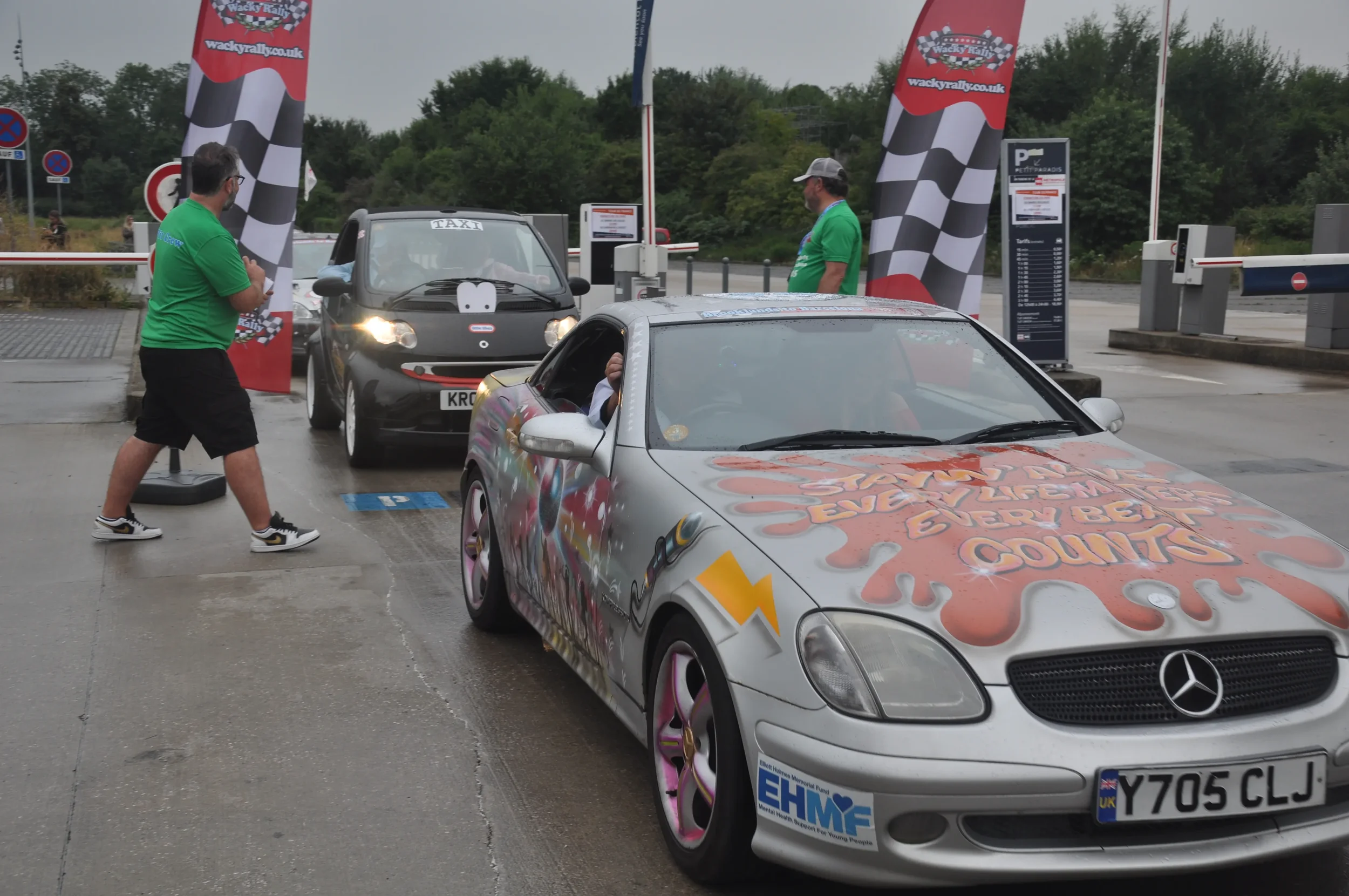 A silver Mercedes-Benz sports car with a colorful painted design and a message that reads 'Start your day with a smile and be thankful for every minute, every hour, every day, every week, every month' on the hood, is near a black smart car with a smi