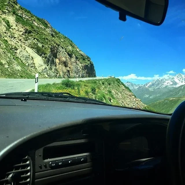 Inside view of a vehicle on a mountain road with a cliff on the left and snow-capped mountains in the distance under a clear blue sky.