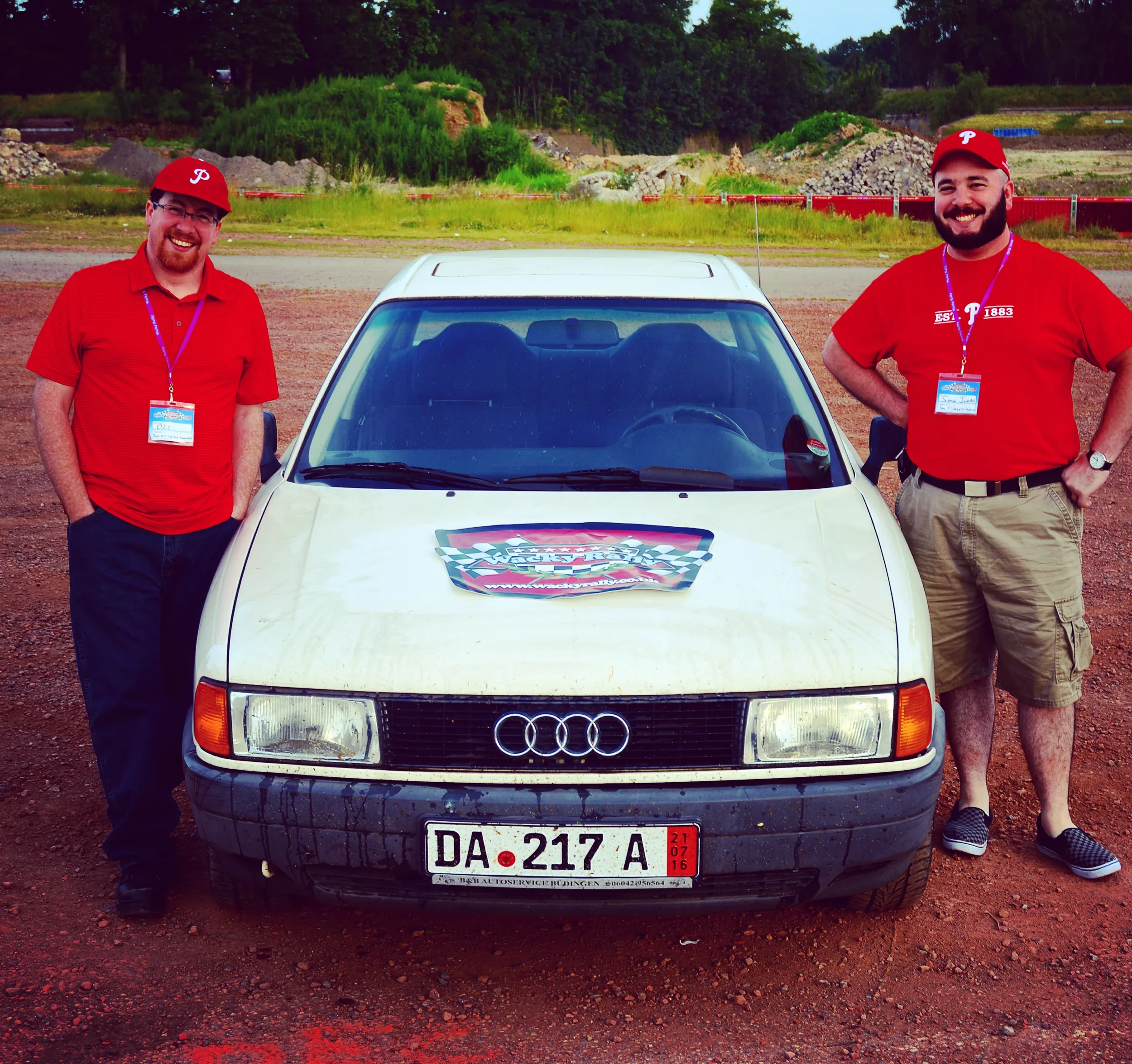 Two men in red shirts and hats stand next to a white Audi with a racing-themed sticker on the hood. They are outdoors on a dirt lot with greenery and hills in the background, smiling at the camera.