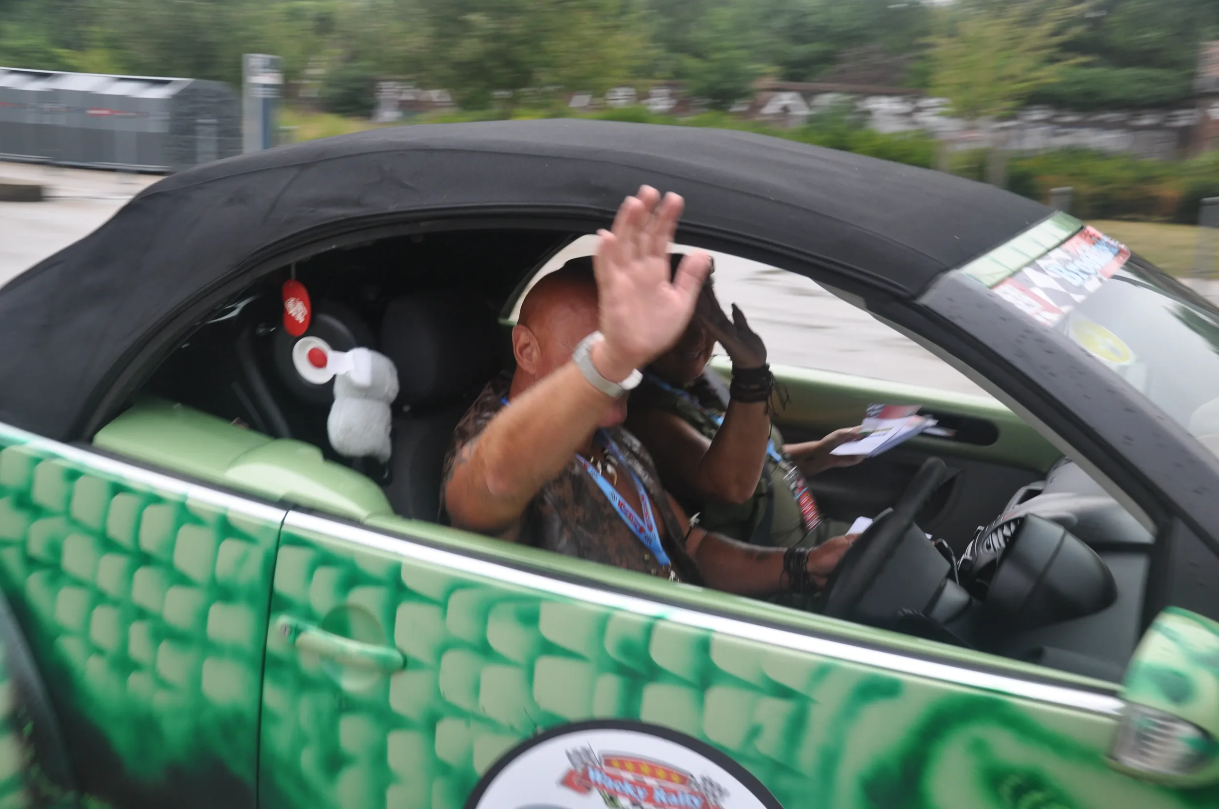 A man in a black shirt is sitting in a small green race car, waving with his left hand while holding papers in his right hand. The car's interior has a plush toy with large headphones attached.