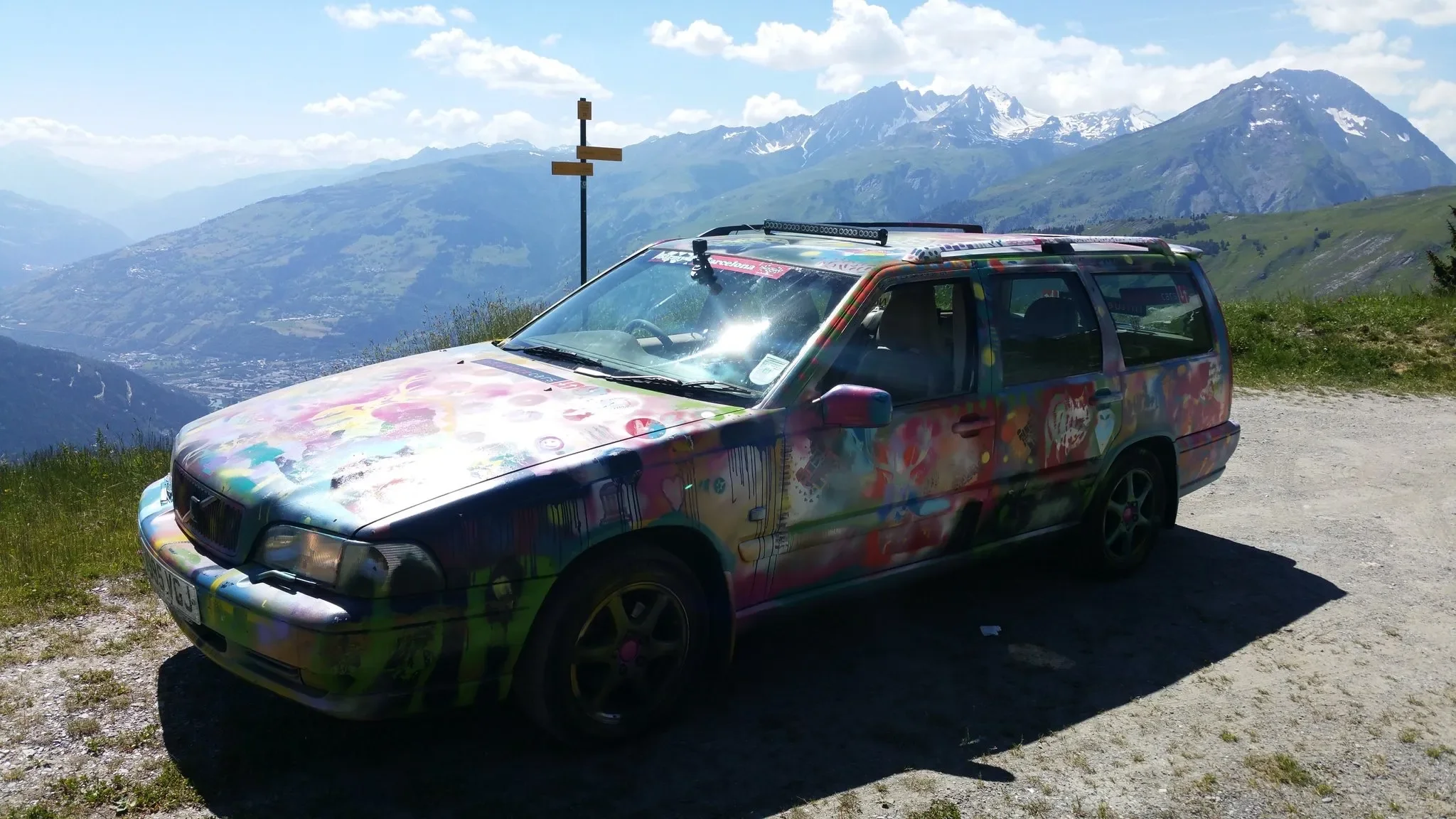 A colorful graffiti-painted station wagon parked on a mountain overlook with snow-capped peaks in the background.