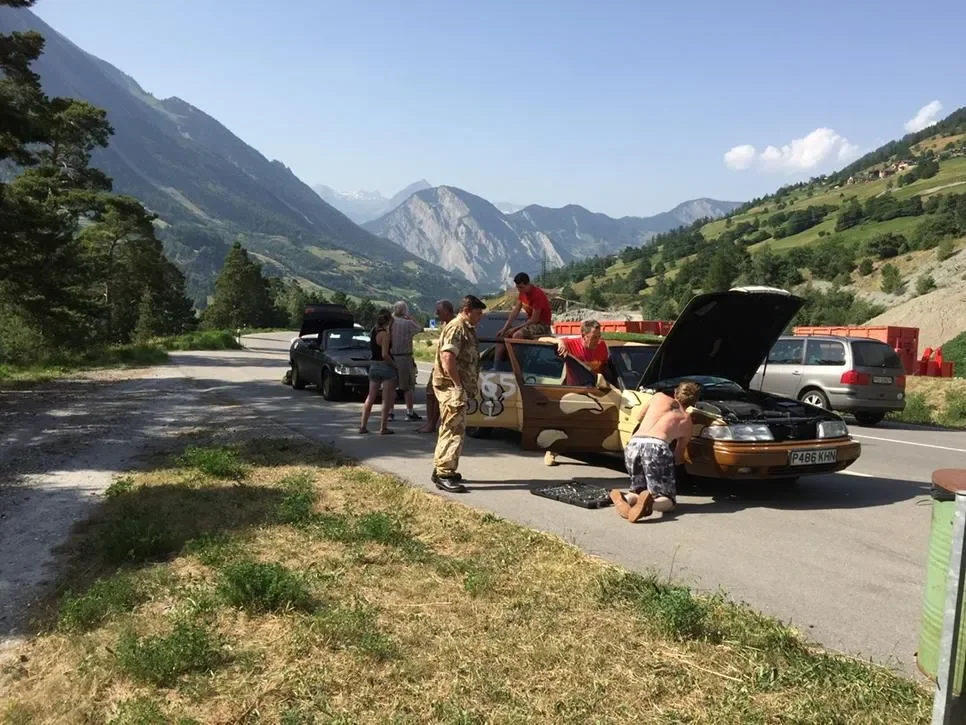 Group of people fixing or inspecting a vintage car on the side of a scenic mountain road in a remote area.