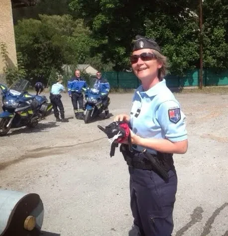 Smiling female police officer holding gloves in an outdoor parking lot, with police motorcycles and other officers in the background.