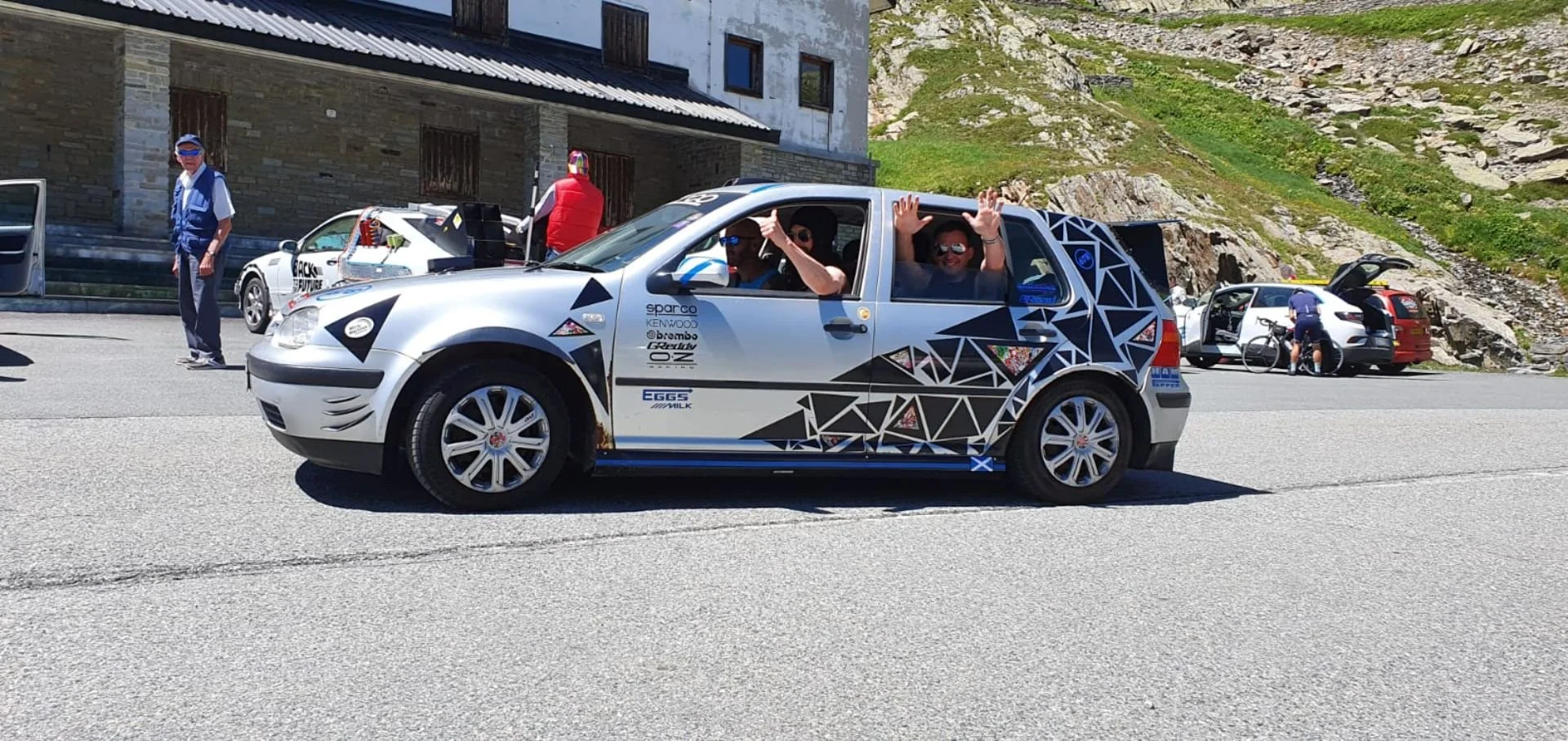 A silver car with geometric designs and sponsor stickers, with two people waving from the driver and passenger seats, parked by a mountain road with other vehicles and a cyclist in the background.