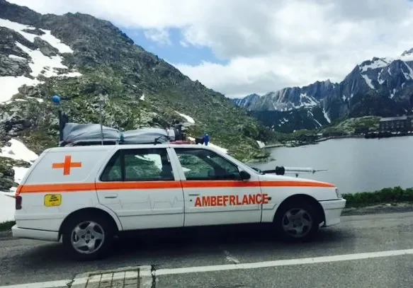 A white ambulance vehicle with an orange stripe and red cross parked on a mountain road by a lake, with snow-capped mountains and green hills in the background.