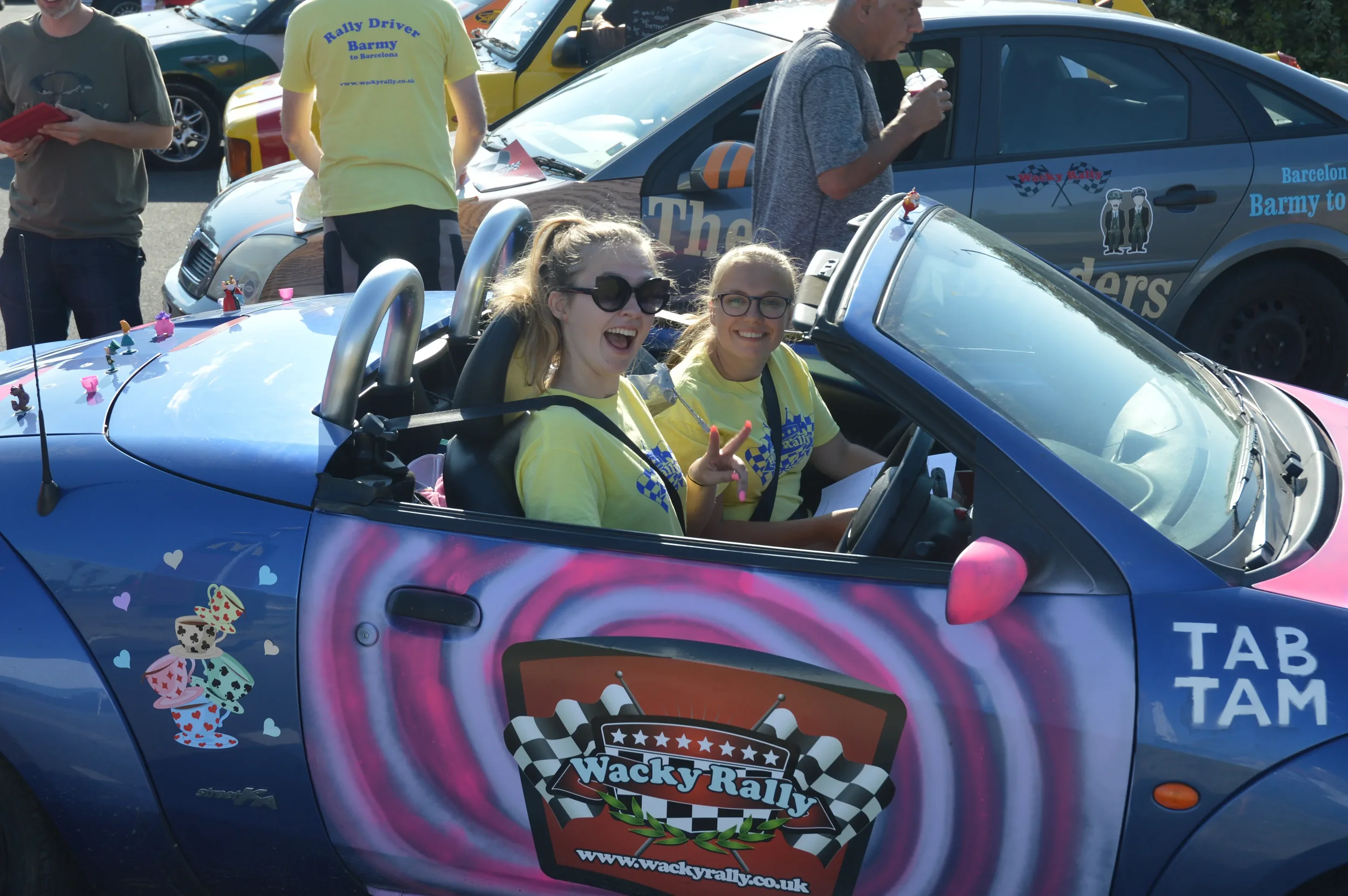 Two young women sitting in a decorated blue convertible car during a rally event, smiling and making peace signs. The car has the 'Wacky Rally' logo on the side, and there are people and other cars in the background.