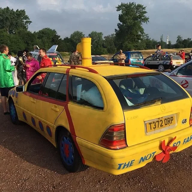 A yellow car designed to look like the Ecto-1 from Ghostbusters, with the phrase "The Not Ecto-Fan" and a red propeller sticker on the back, parked at an outdoor event with other vehicles and people in the background.