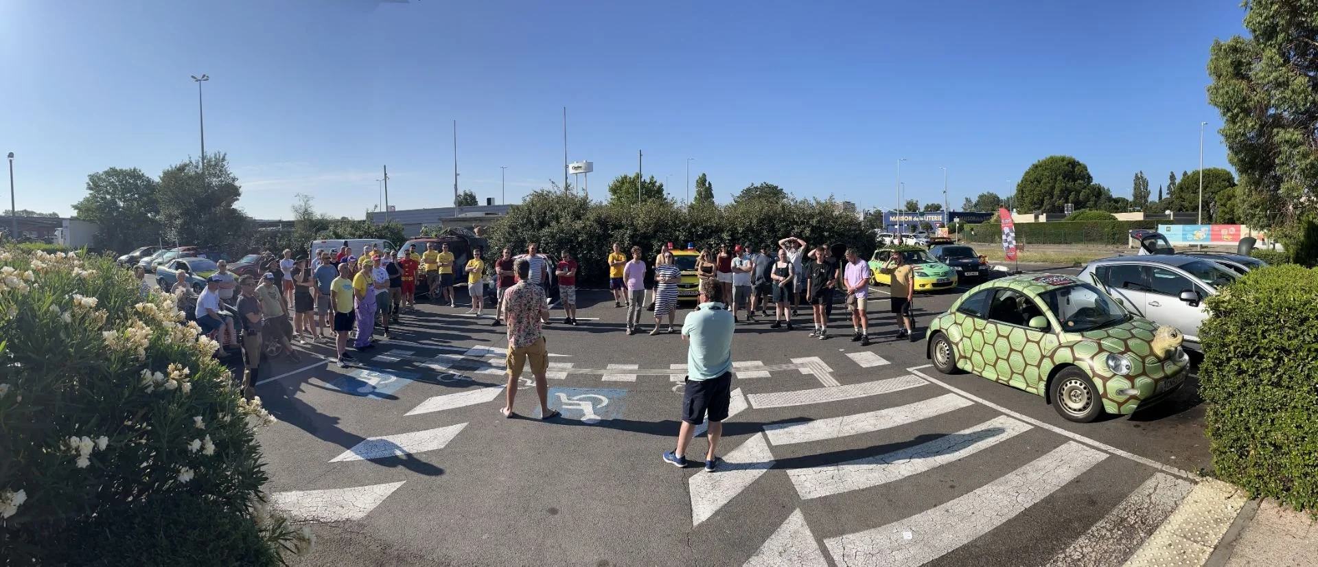 A large group of people gathered in a parking lot on a sunny day, with some cars visible, including a small car painted with a turtle shell pattern. The crowd appears to be attending an event, with some individuals wearing brightly colored clothing a