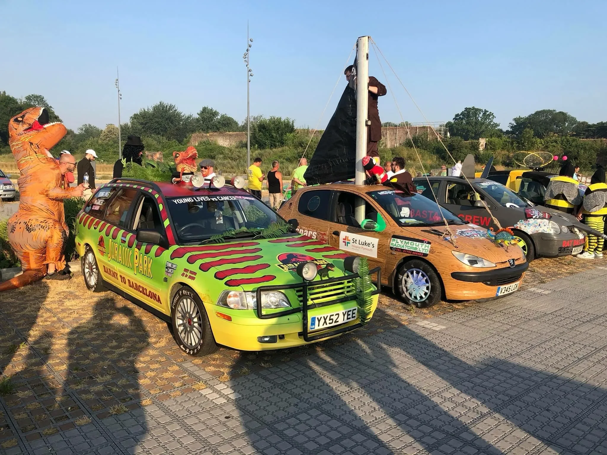 Car decorated as a dinosaur with people in dinosaur costumes and a makeshift boat on top, resembling a Jurassic Park theme at a parade or festival.