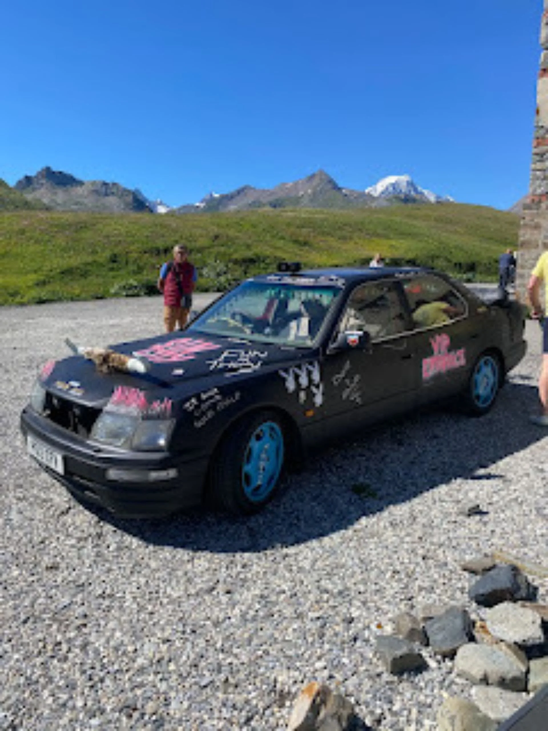 A black car with pink and white decals on a gravel area in a mountain landscape. There are a few people around, and tall peaks with snow in the background.