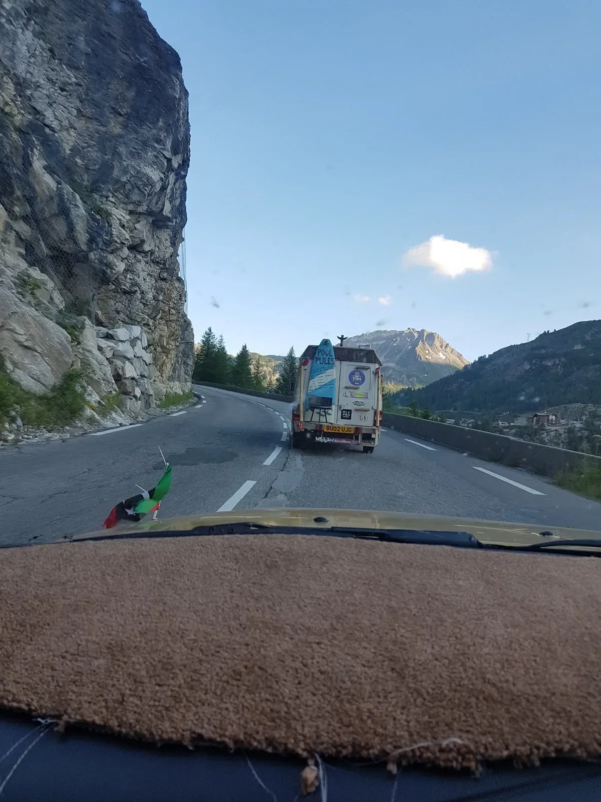 A mountain road with a vehicle in front, carrying pool supplies, and a small Italian flag on the dashboard inside the car.