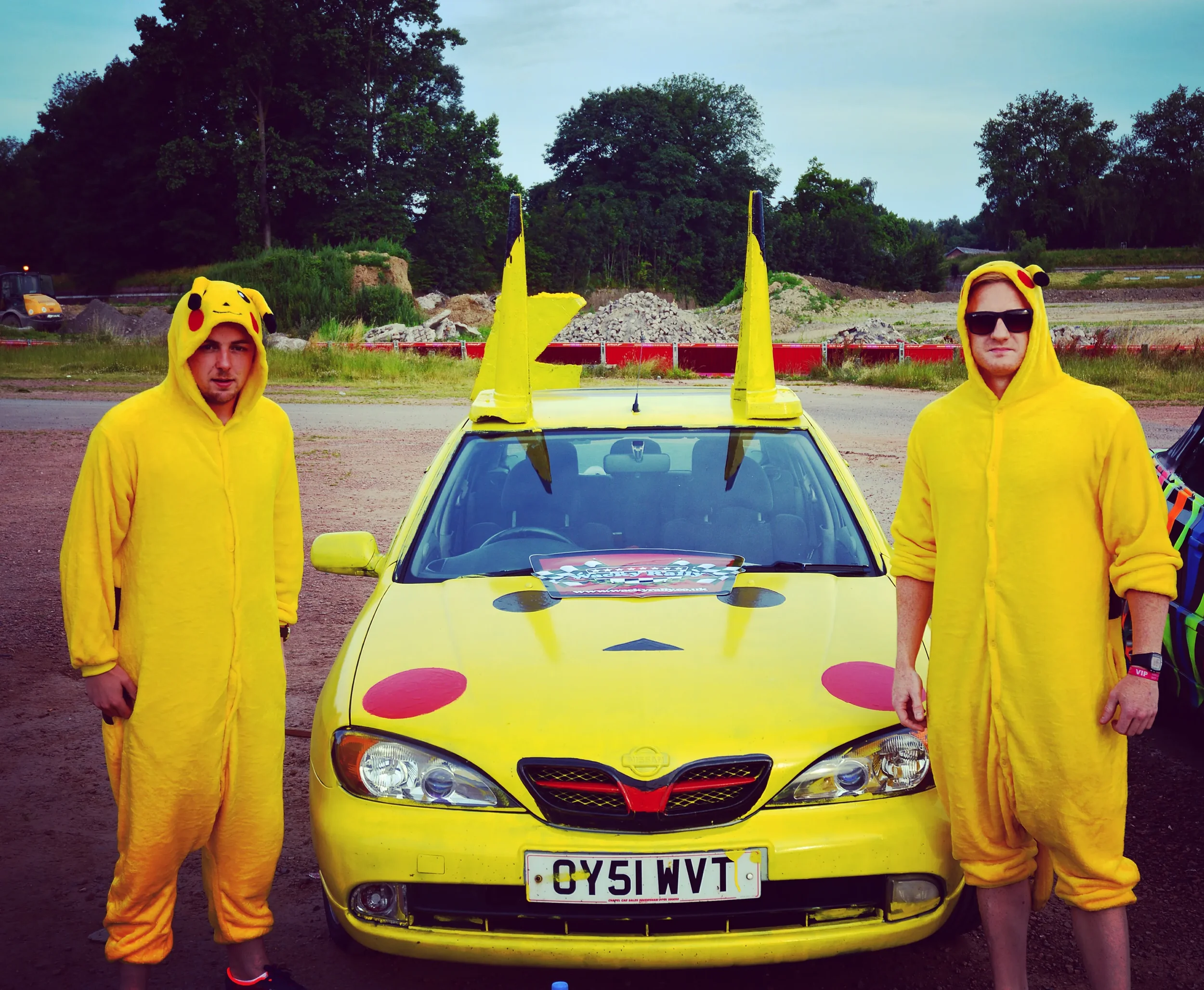 Two young men dressed in yellow Pikachu onesies stand on either side of a yellow car decorated with Pikachu-themed features, including Pikachu ears on the roof and large red cheeks on the hood, during daytime outdoors.