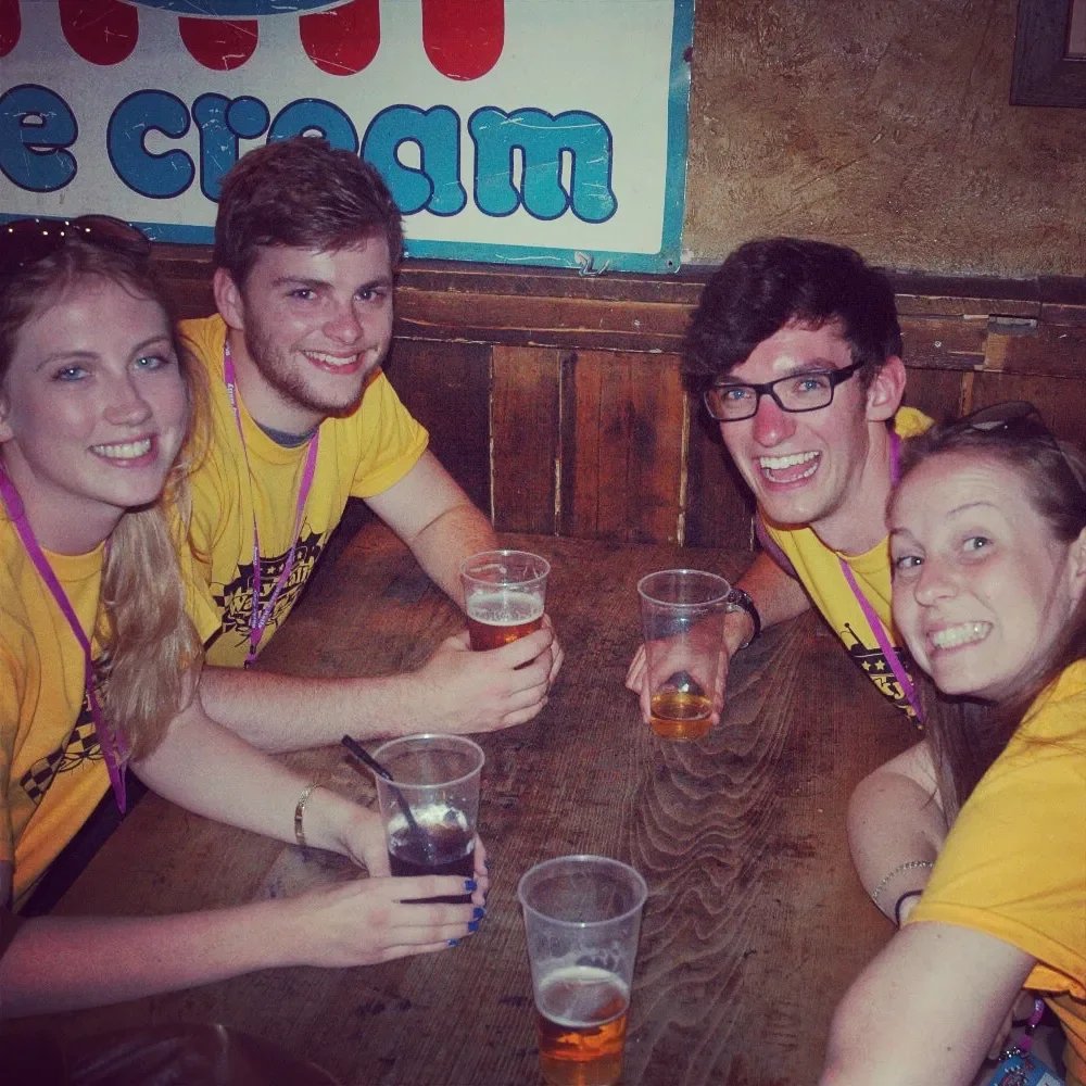 Four young adults sitting at a wooden table in a bar, smiling, with drinks in plastic cups, wearing yellow shirts, and purple lanyards, in a lively indoor setting.