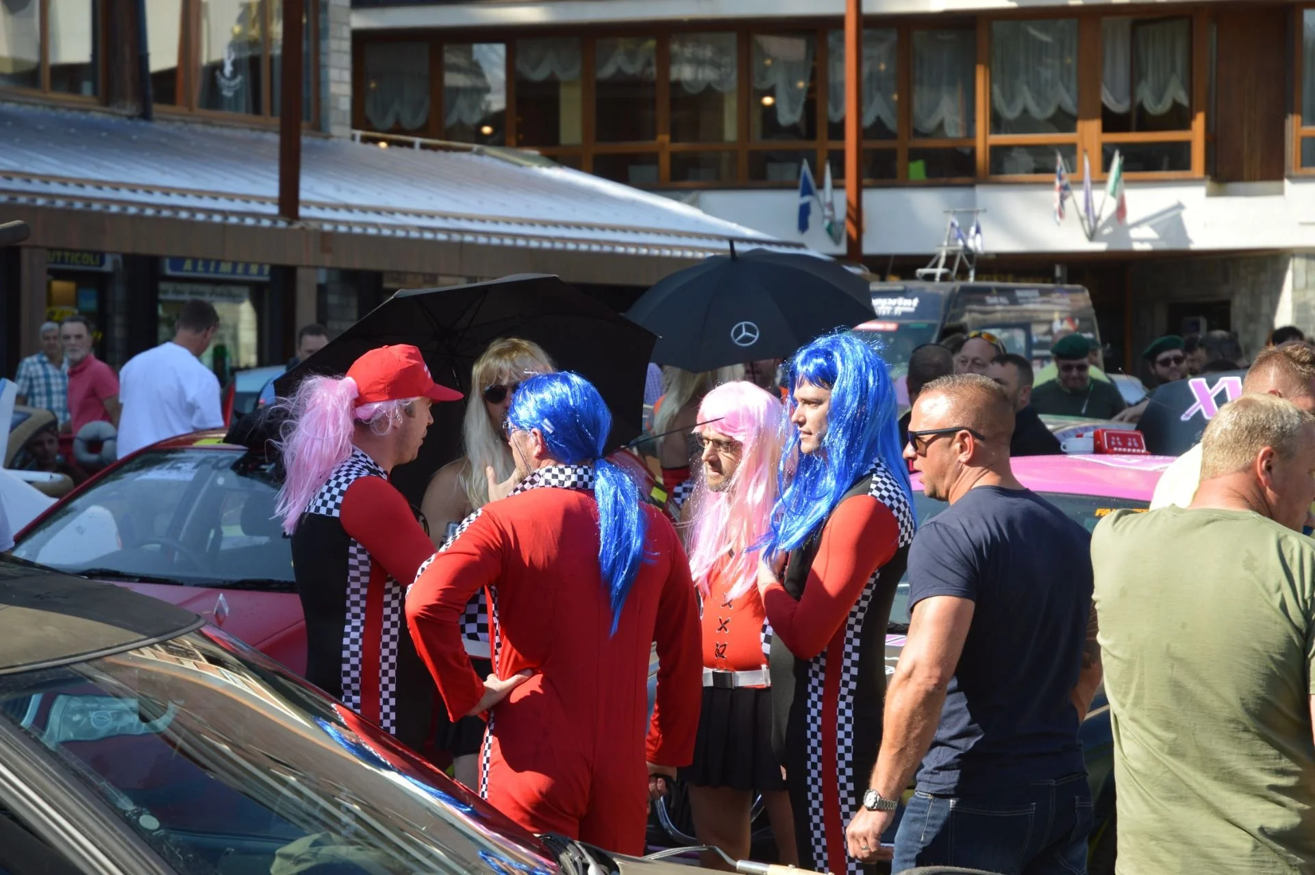 People dressed in racing-themed costumes with colorful wigs, standing among vintage cars in a crowded outdoor setting.